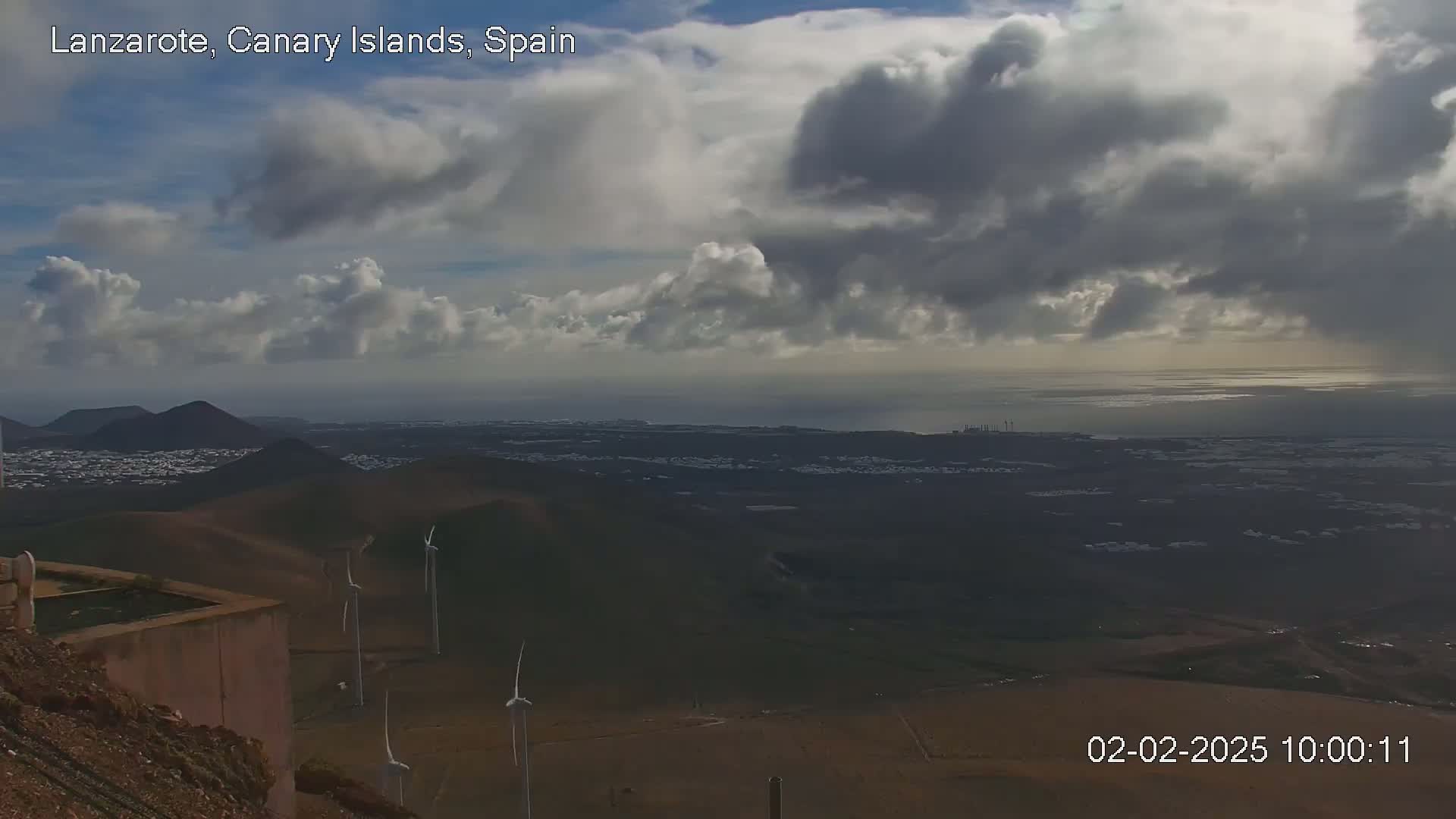 A panoramic view of a volcanic landscape with wind turbines, a town, and ocean under a partly cloudy sky.