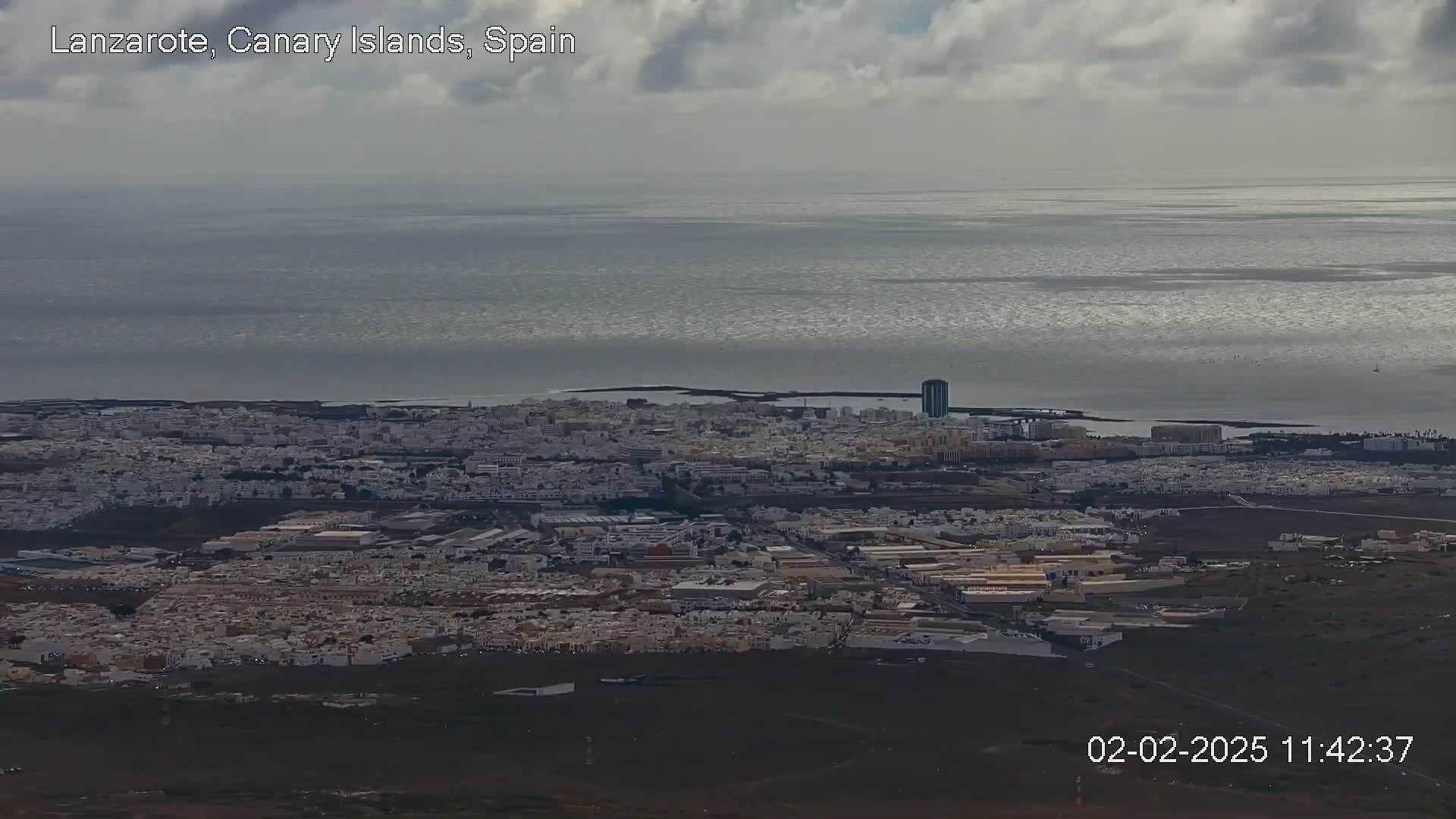 A coastal city with low-rise buildings stretches along a calm, gray ocean under a cloudy sky.