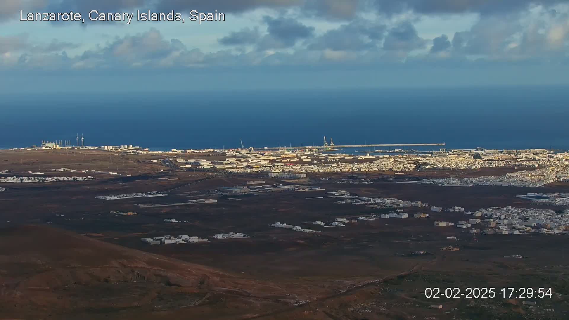 A high-angle view of a coastal town under a partly cloudy sky, showing a harbor and ocean in the distance.