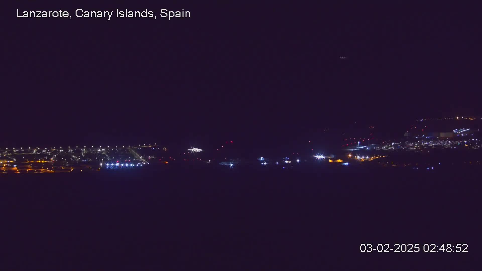 A nighttime long shot shows a brightly lit airport at night under a clear, dark sky.