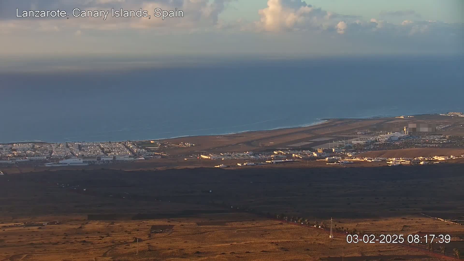 A high-angle view shows a coastal town and airport next to a calm ocean under a partly cloudy sky.