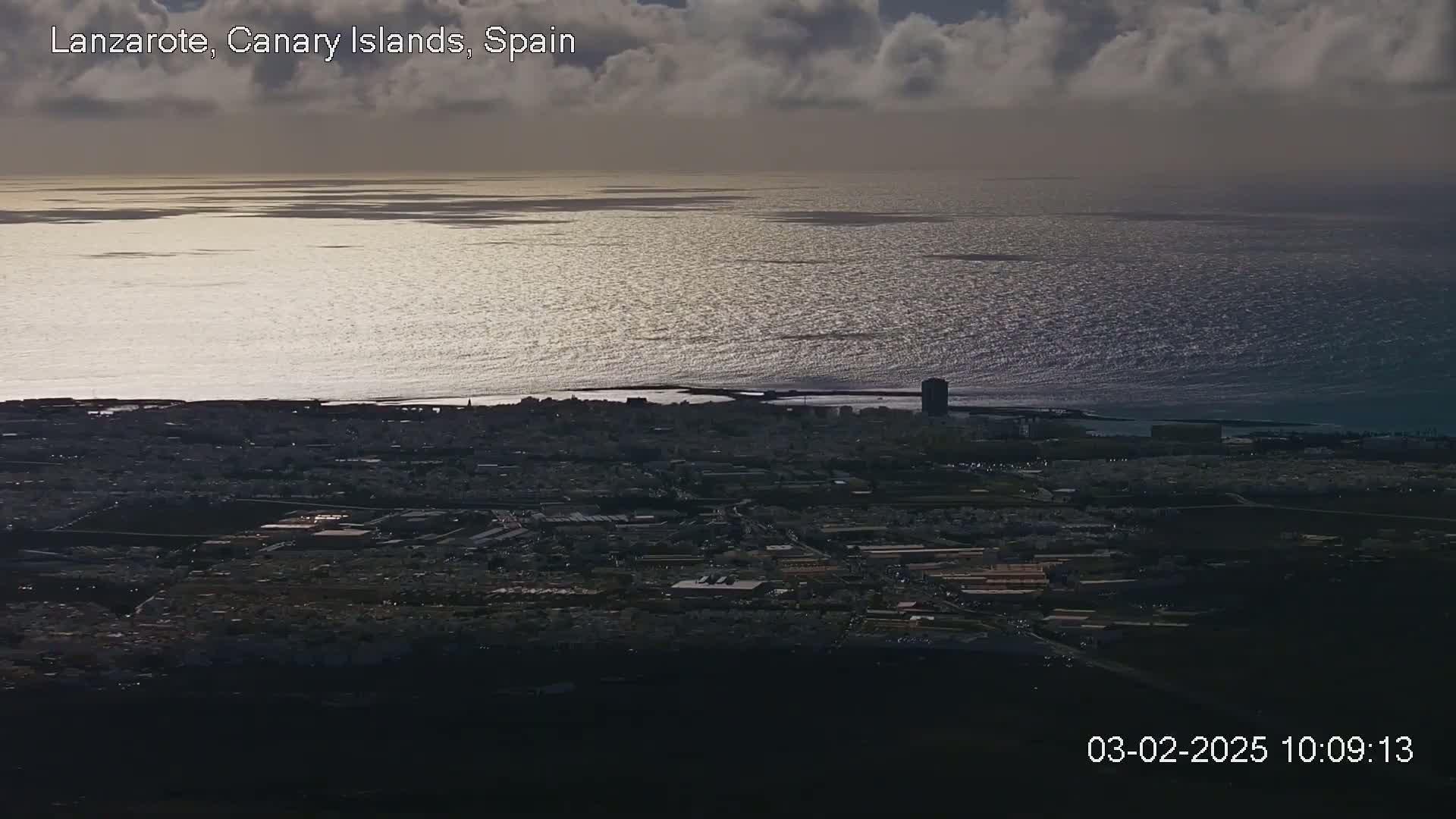 A coastal city is viewed from above under a partly cloudy sky, with a glistening ocean stretching to the horizon.