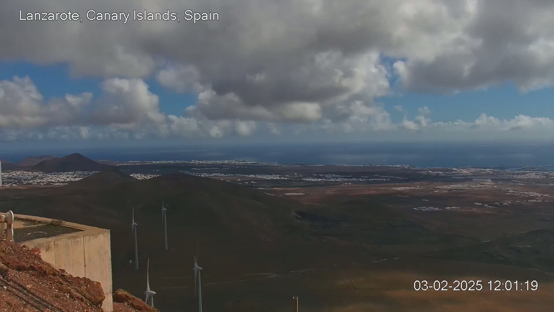 A partly cloudy day reveals a panoramic view of a volcanic landscape with wind turbines and a distant ocean.