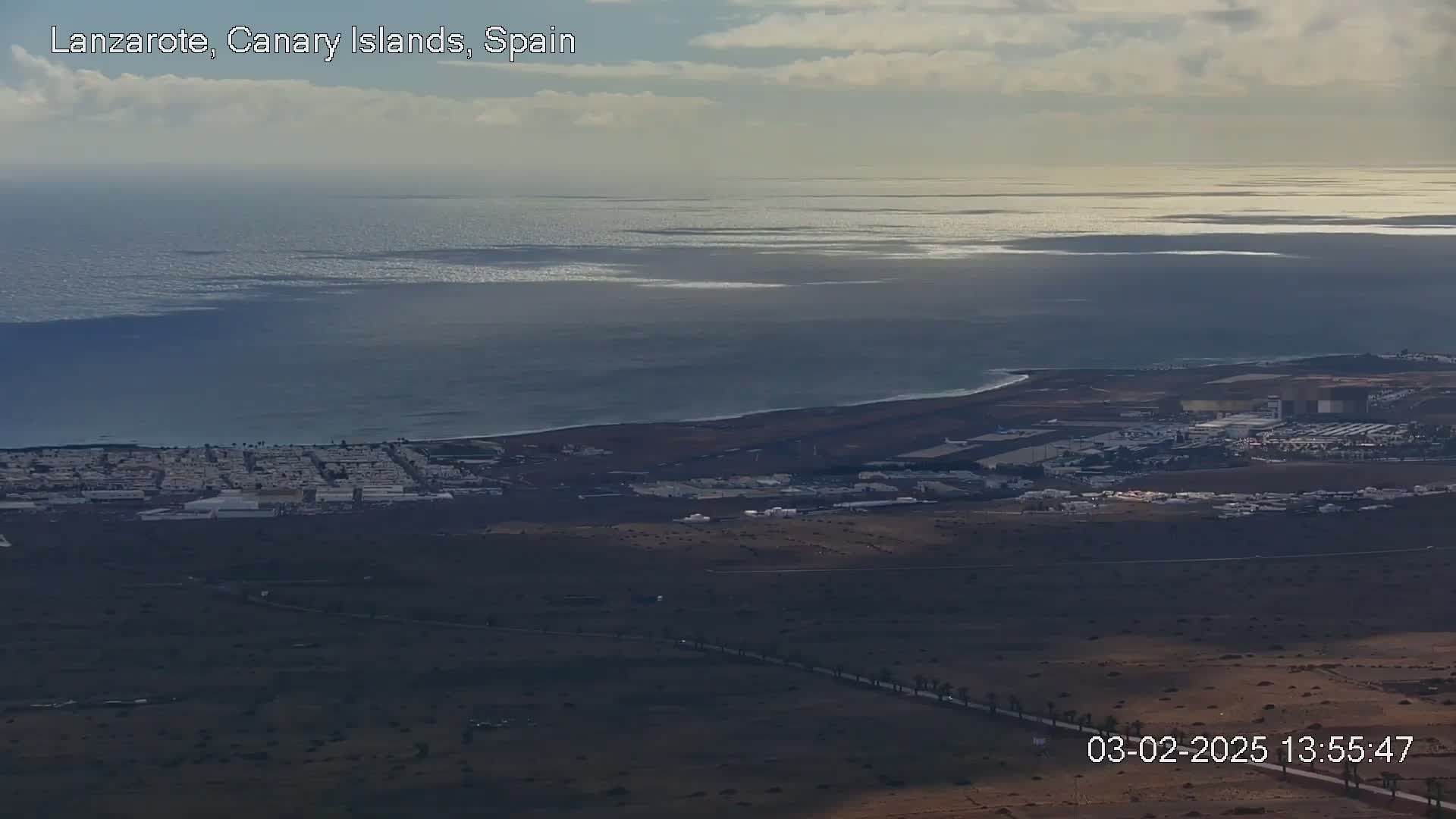 A coastal town with an airport is visible from an elevated viewpoint under partly cloudy skies, overlooking a vast ocean.