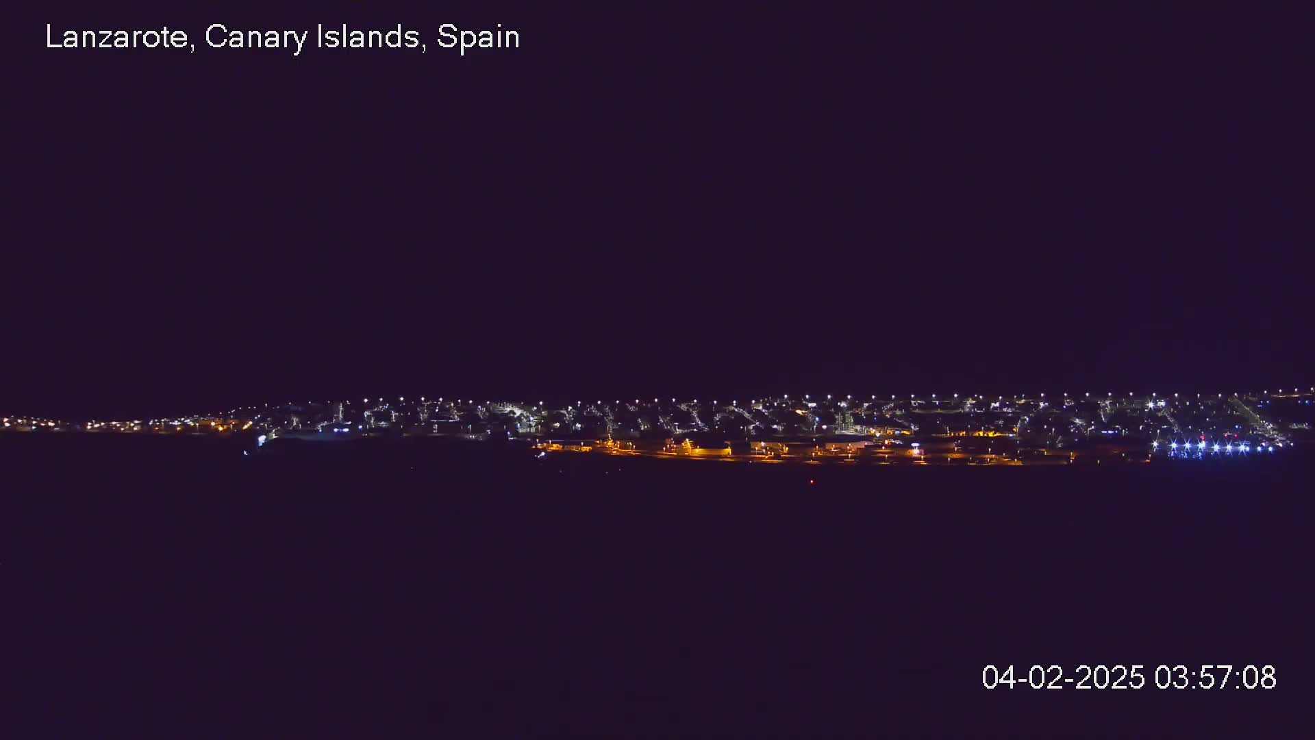 A nighttime long shot shows a town's lights spread across a dark landscape under a clear, dark sky.