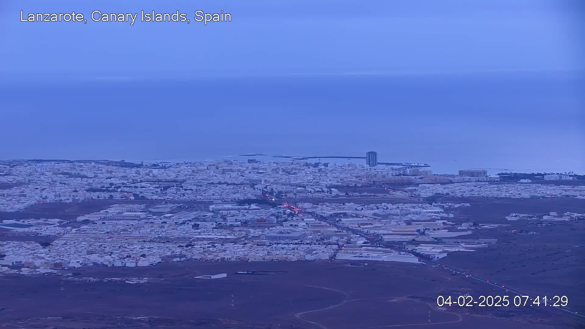 A sprawling coastal city with low-rise buildings extends to a calm ocean under a twilight sky.