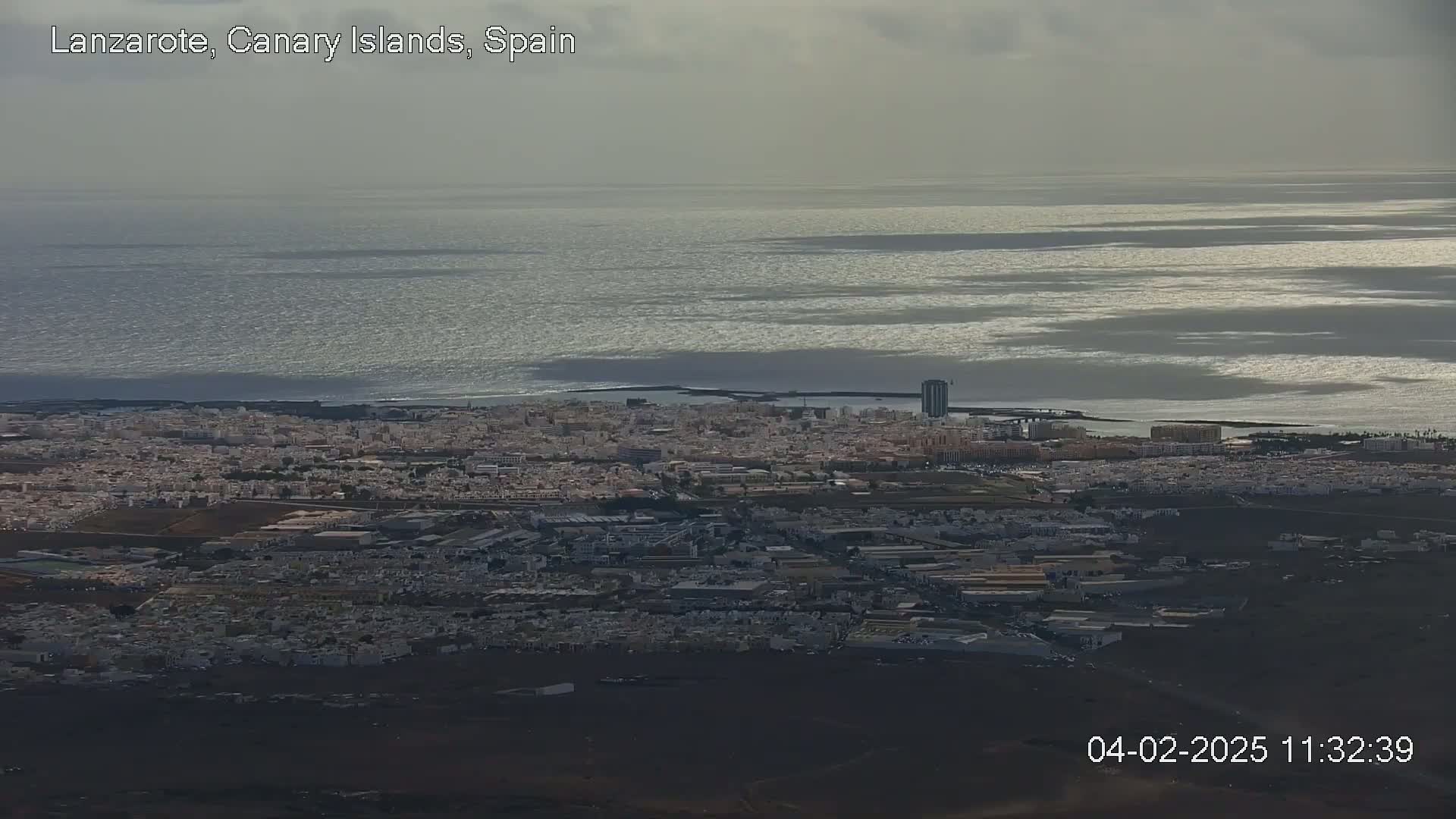 An aerial view shows a coastal city under an overcast sky, with a calm, glittering ocean extending to the horizon.