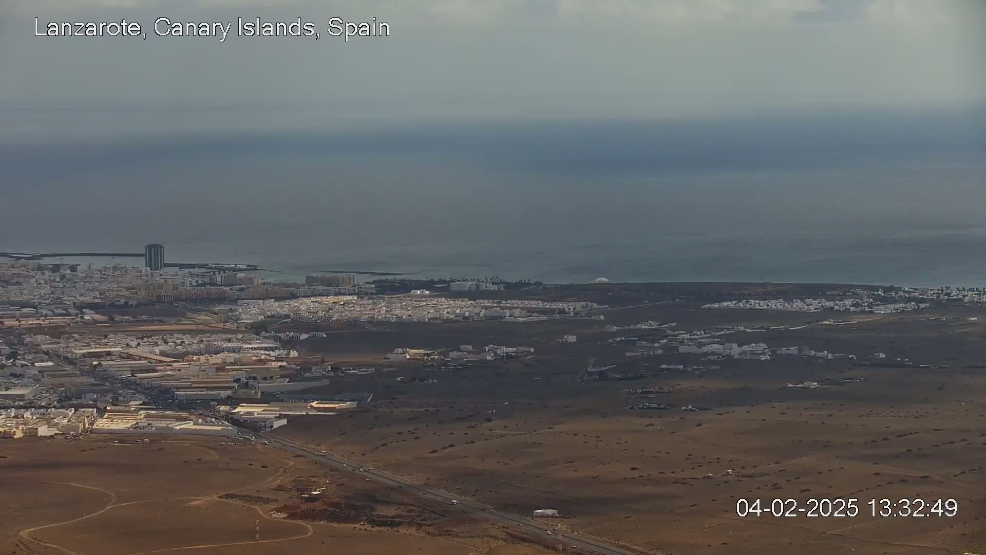 An aerial view shows a coastal town with low, light-colored buildings stretching across a brown, arid landscape that meets a calm, gray-blue ocean under an overcast sky.