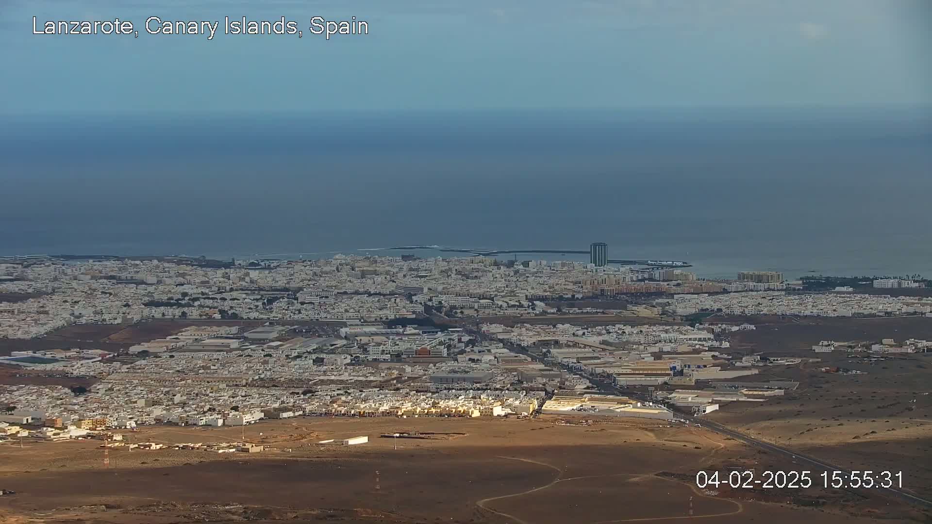 A sprawling coastal city with light-colored buildings extends to a calm ocean under a clear sky.