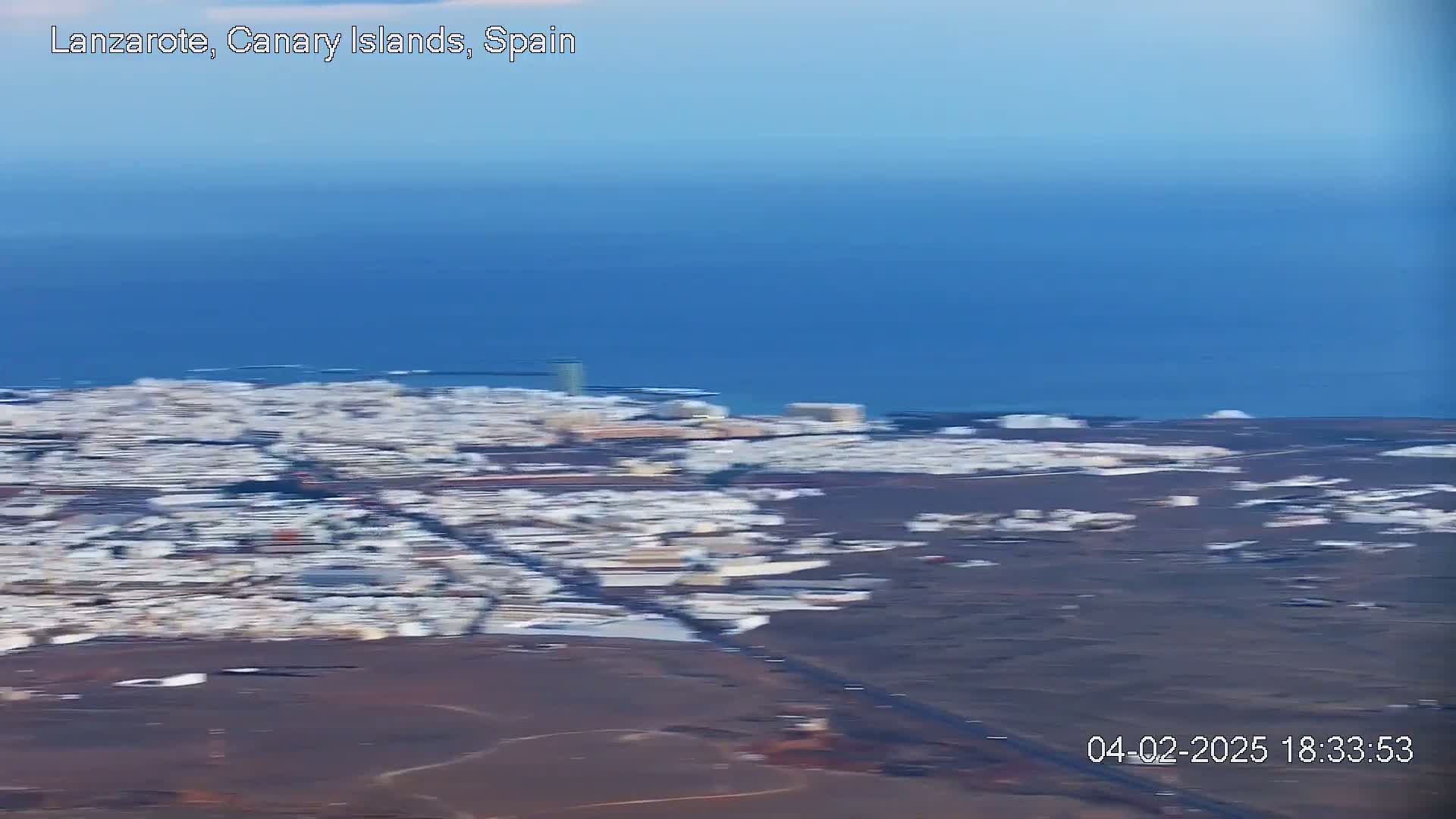 An aerial view shows a coastal town with white buildings against a dark brown landscape, next to a calm blue ocean under a clear sky.