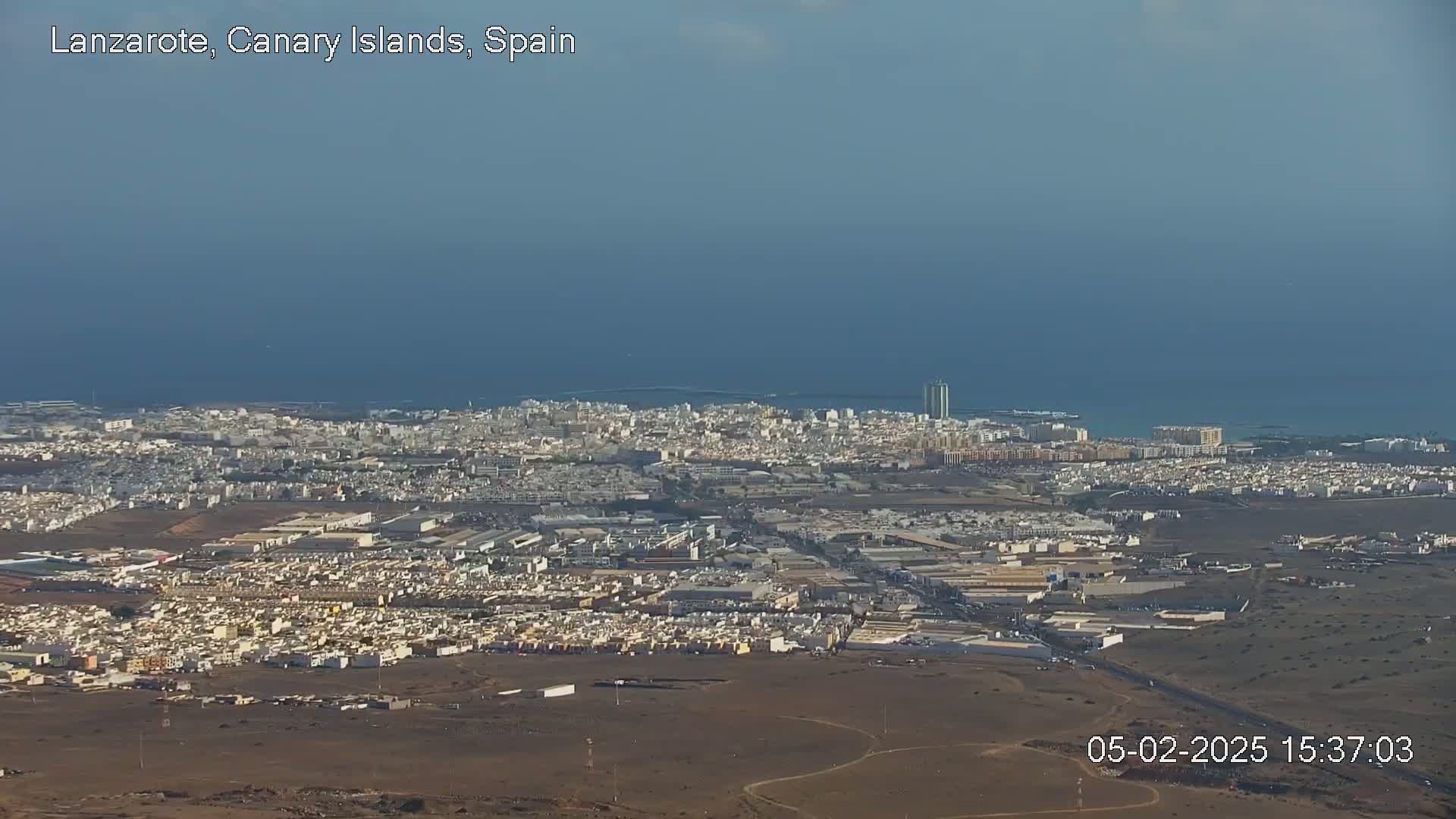A sprawling coastal city with light-colored buildings extends to a calm ocean under a clear, light-blue sky.