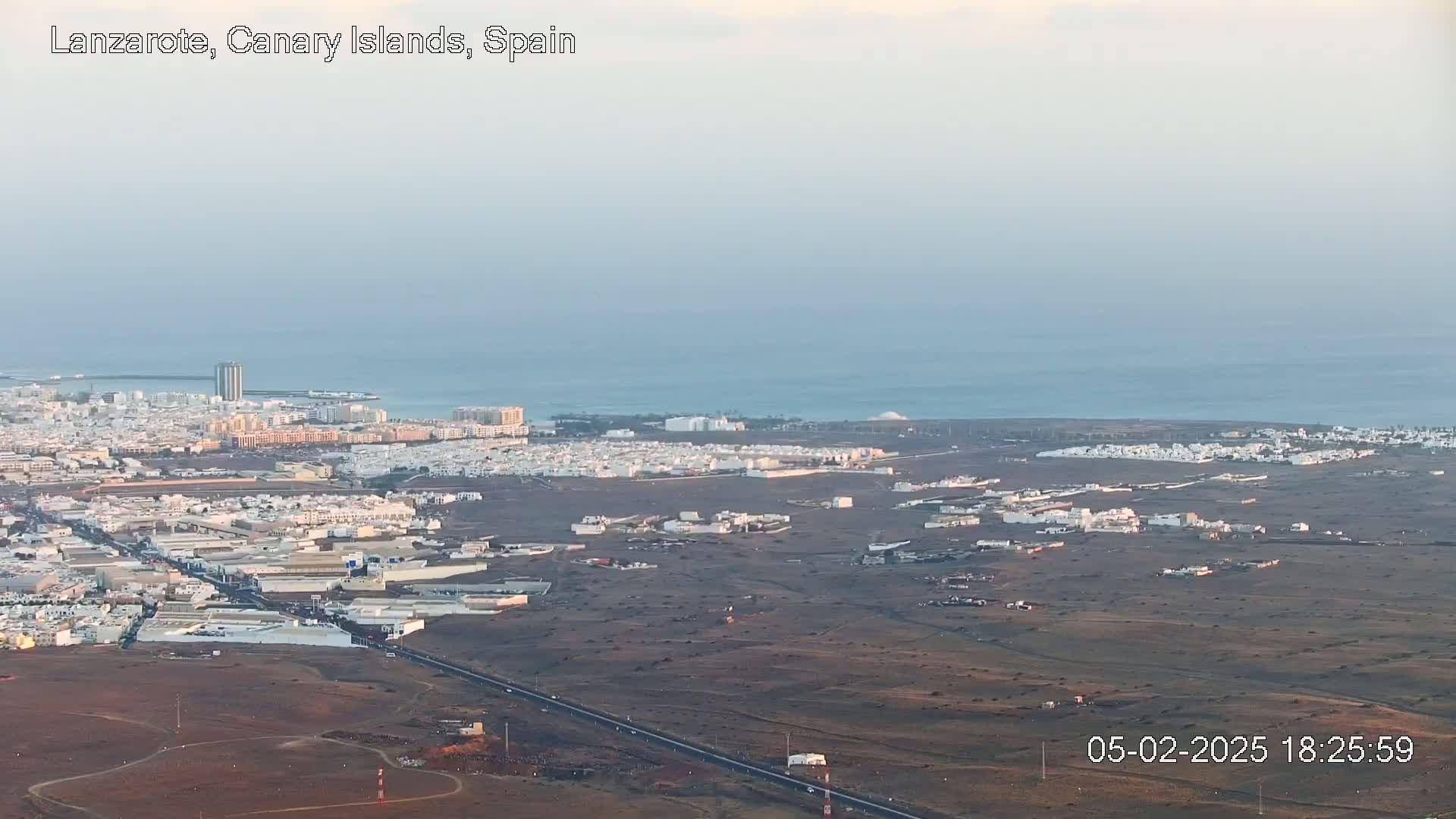 An aerial view shows a coastal city with light-colored buildings and a calm ocean under a hazy sky.