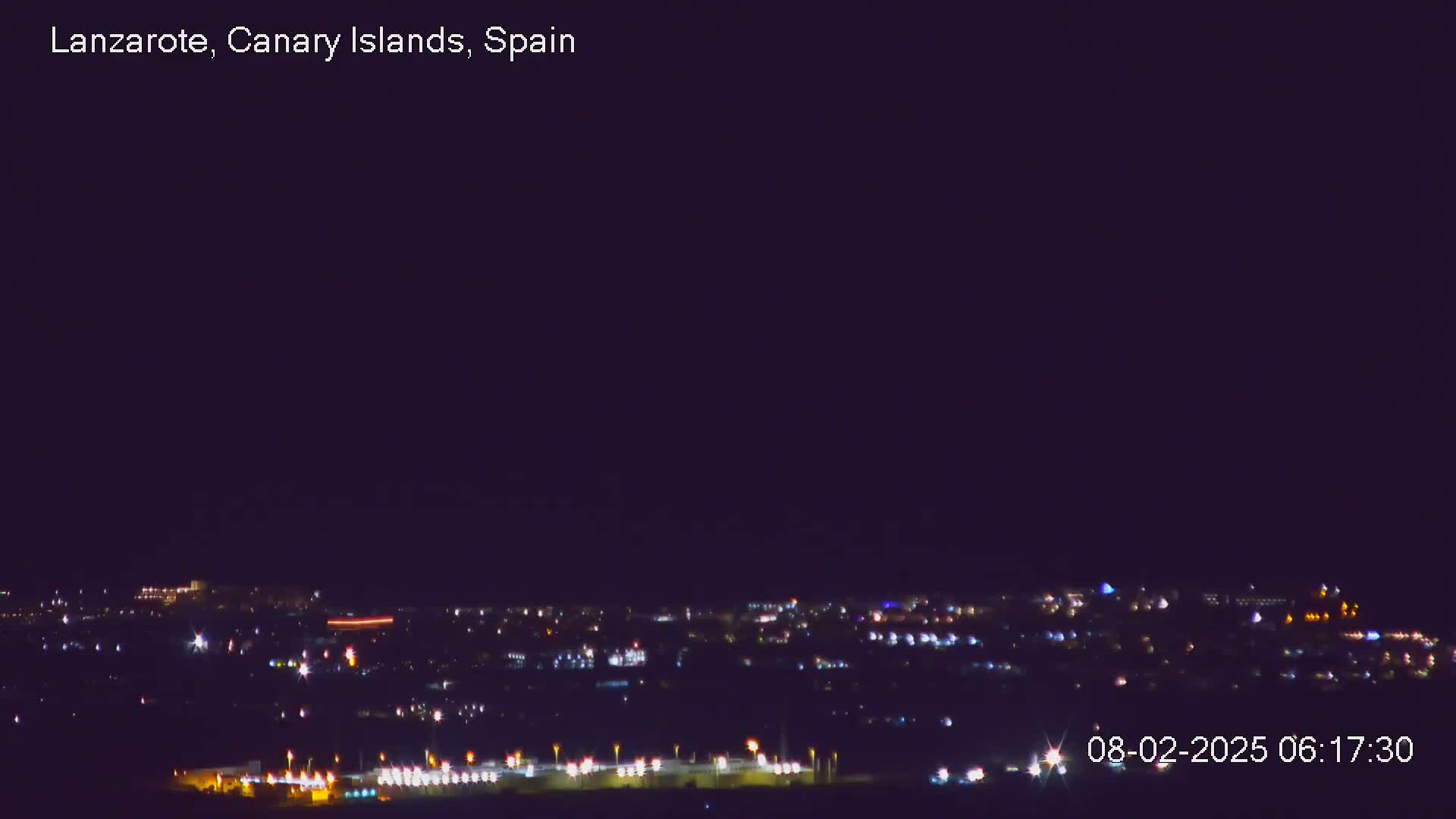 A nighttime long shot shows a city's lights spread across a dark landscape under a clear, dark sky.