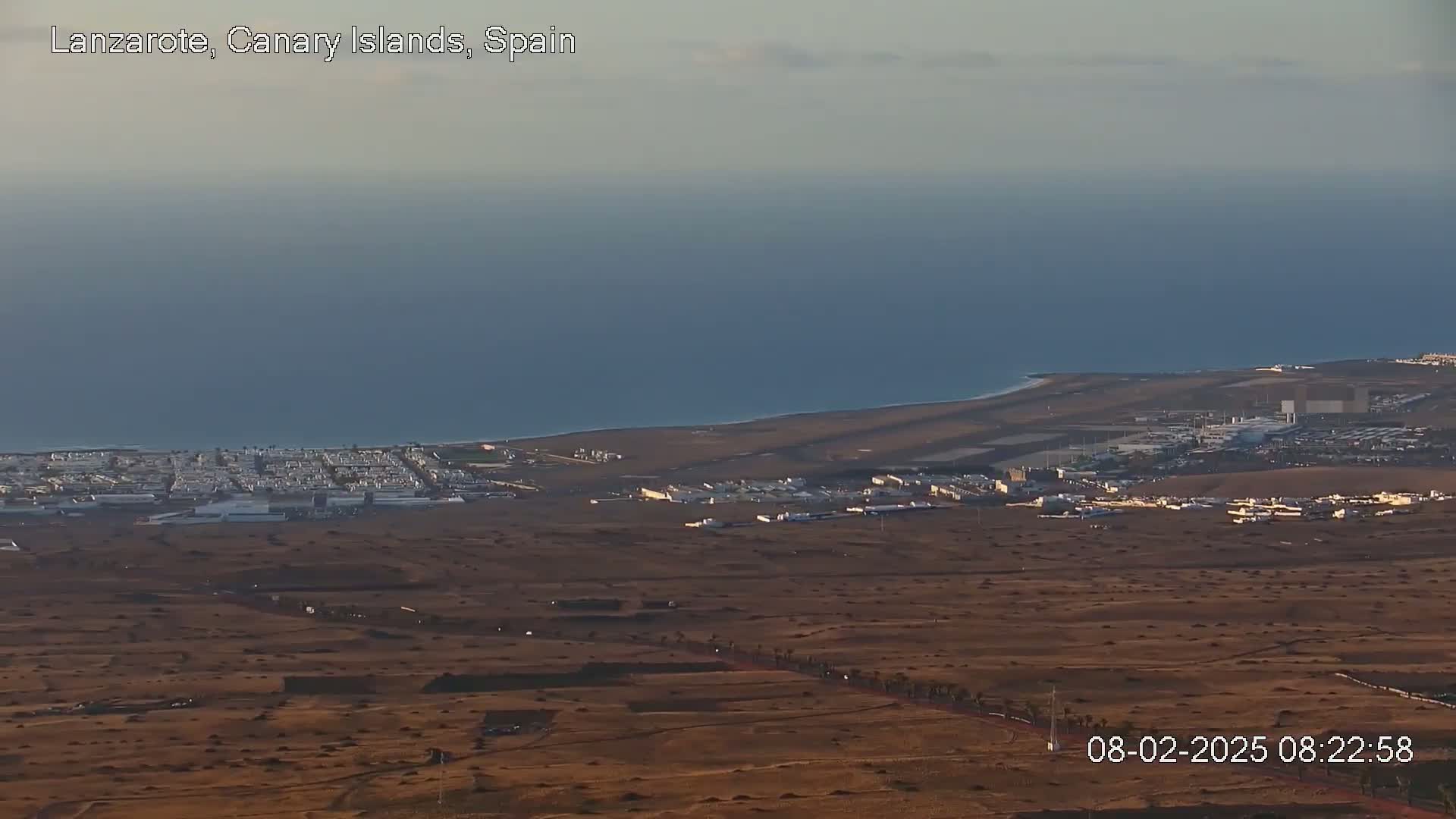 A coastal town and airport are situated on a flat, arid landscape next to a calm ocean under a hazy sky.