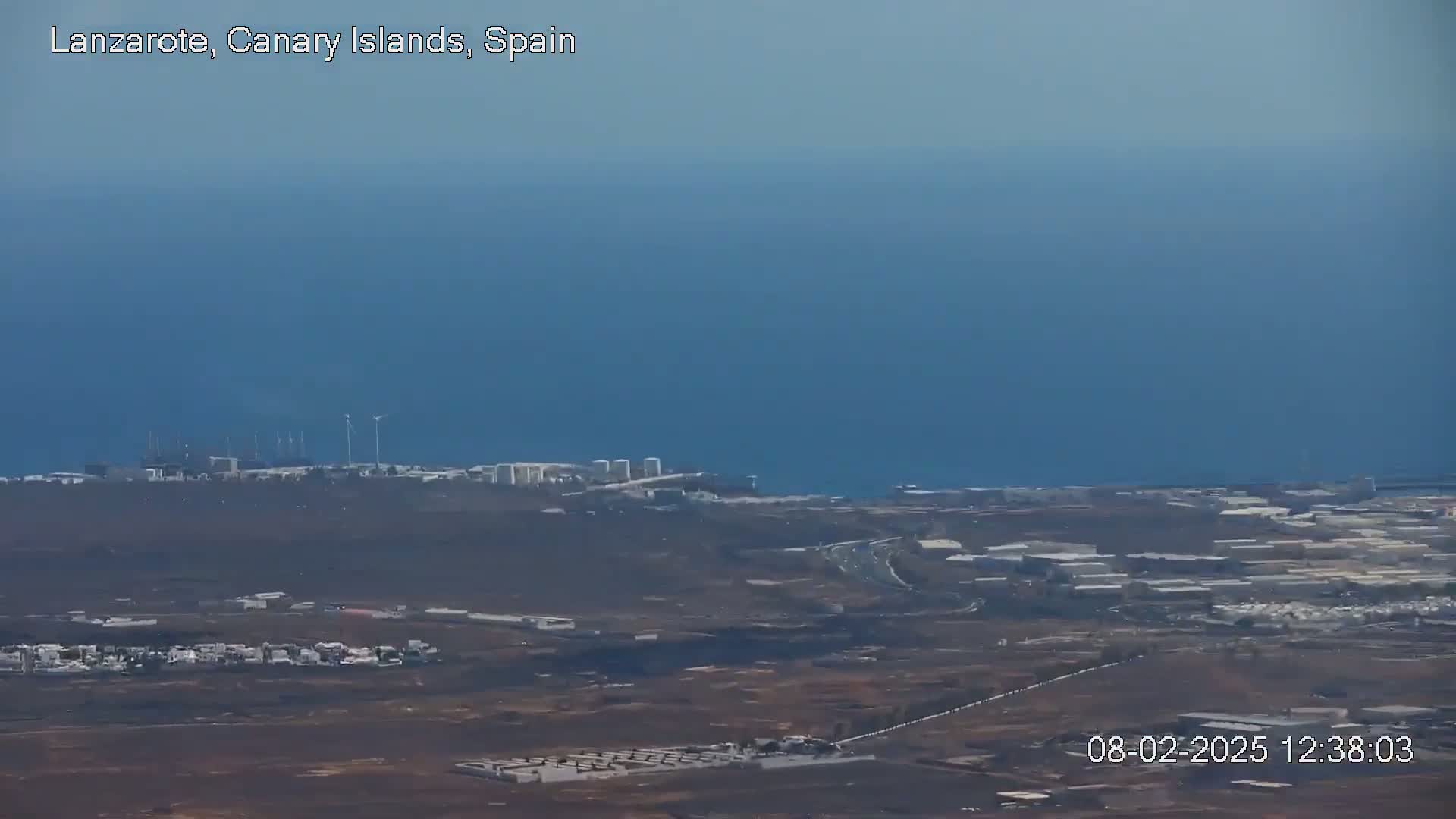 A coastal industrial area with wind turbines and a body of water under a clear sky is seen from a high vantage point.