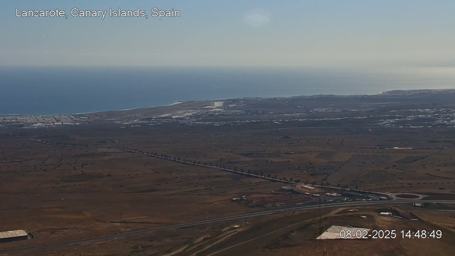 A high-angle, long shot shows a coastal area with a flat, dry landscape, a road lined with palm trees, a small town, and a body of water under a clear sky.