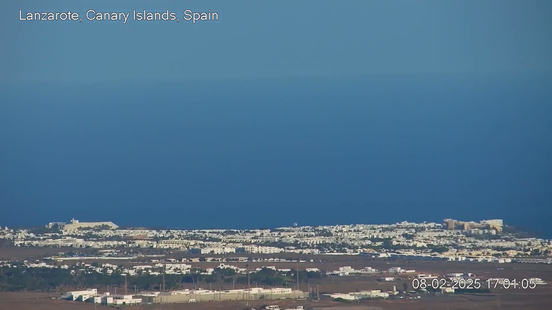 A sprawling coastal town with low-rise white buildings extends to a calm, deep blue ocean under a clear sky.