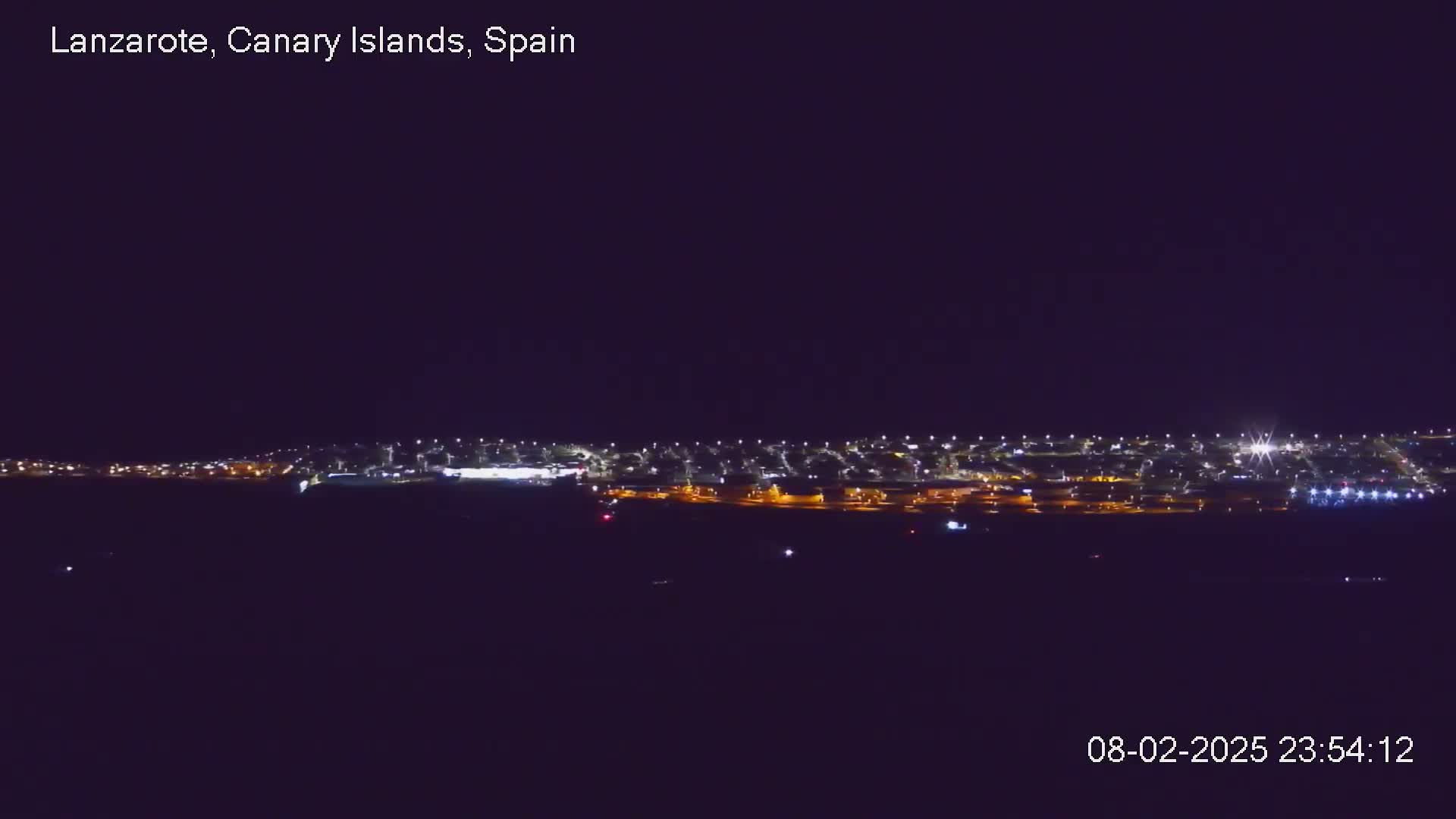 A nighttime long shot shows a brightly lit town or city under a clear, dark sky.