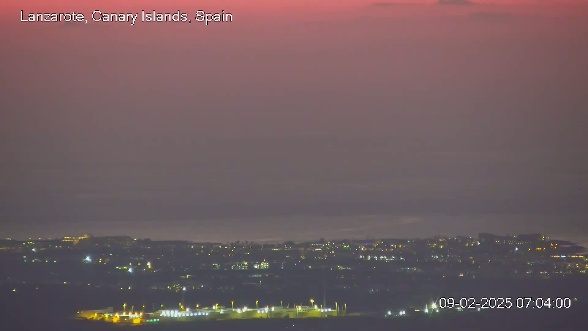 A hazy, pinkish-purple twilight sky overlooks a coastal town illuminated by scattered lights at night.