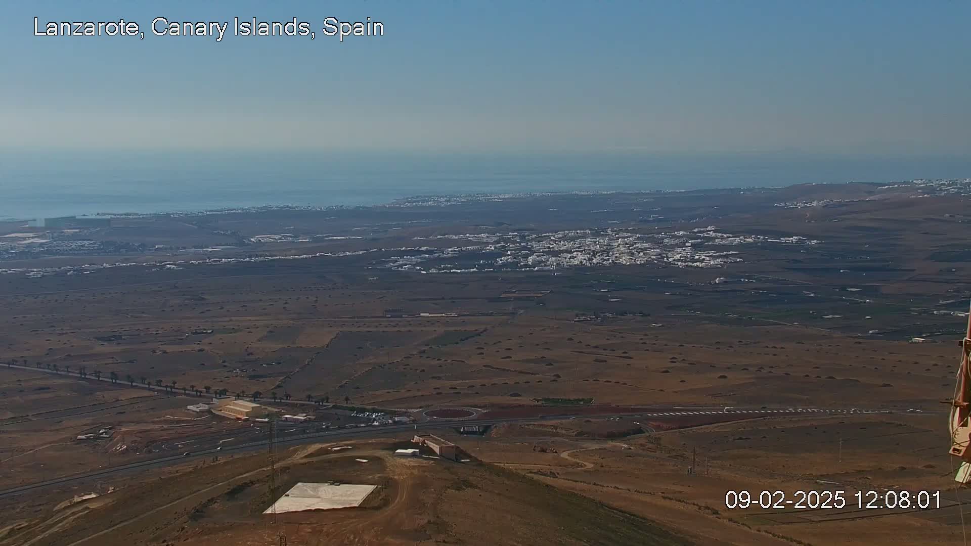 A high-angle, long shot shows a vast, dry, brown landscape with a town and ocean in the distance under clear skies.