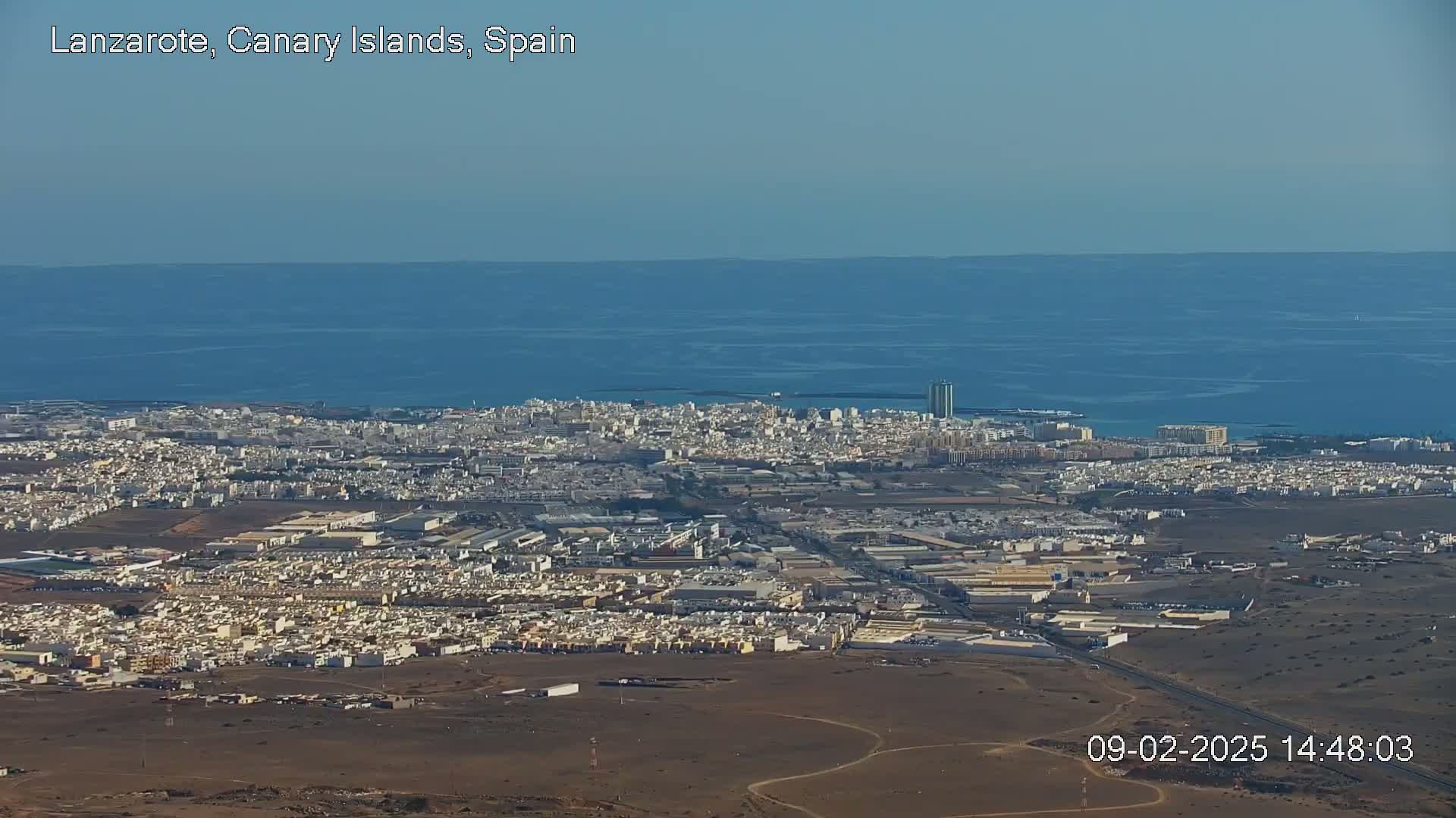 A sprawling coastal city with light-colored buildings extends to a calm, blue ocean under a clear sky.