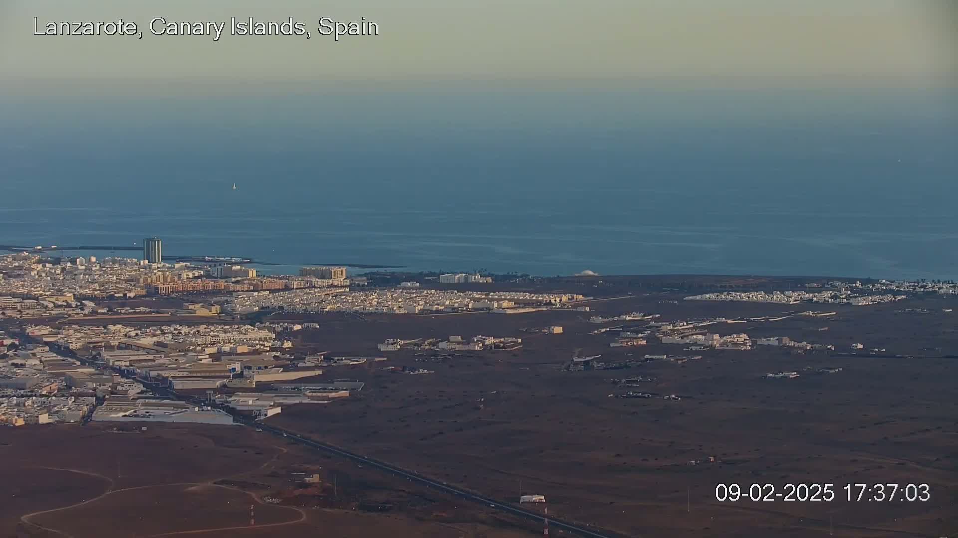 An aerial view shows a coastal town with light-colored buildings sprawling across a dark brown landscape, next to a calm, dark blue ocean under a clear sky.