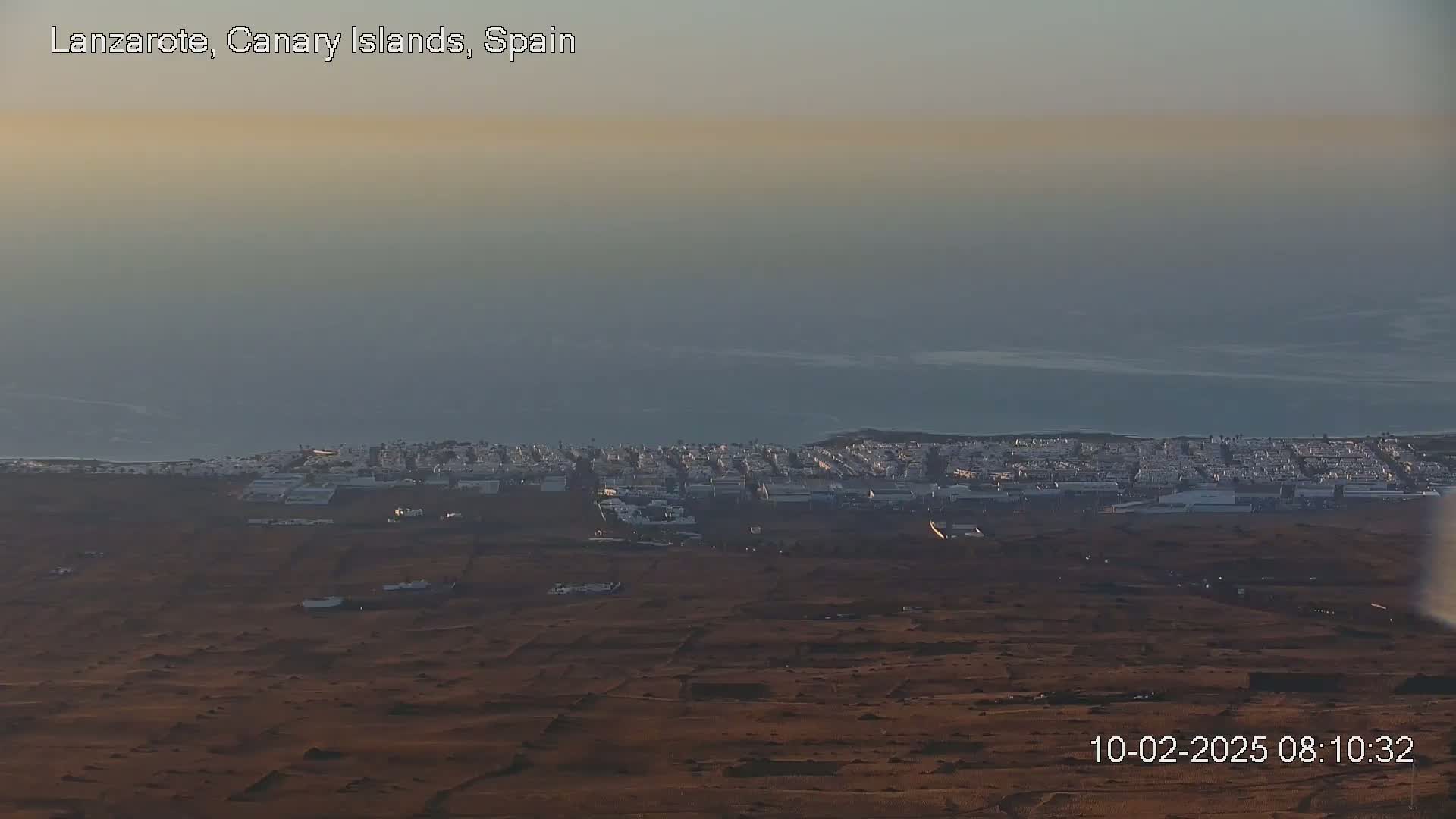 A coastal town with light-colored buildings stretches along a dark brown, arid landscape under a hazy, pale sky.