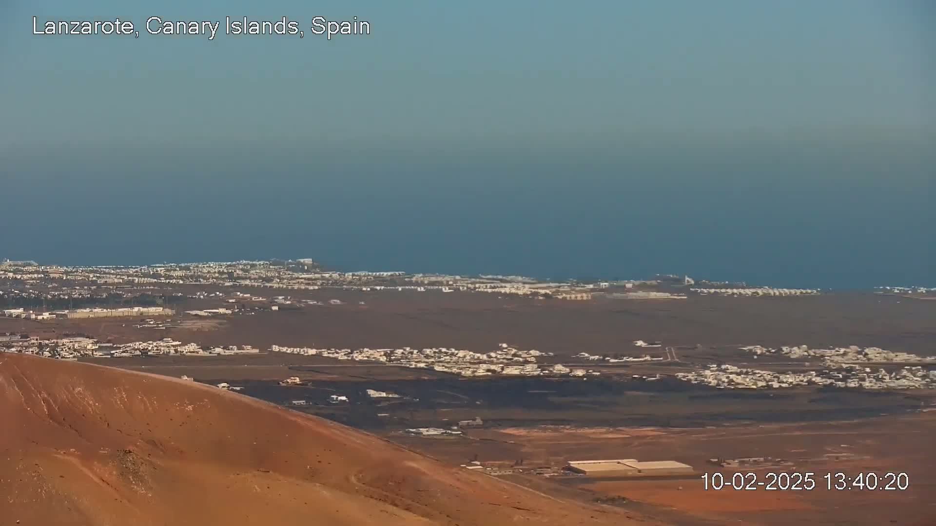A sprawling coastal town and ocean are viewed from a high vantage point on a clear day, with a reddish-brown hill in the foreground.