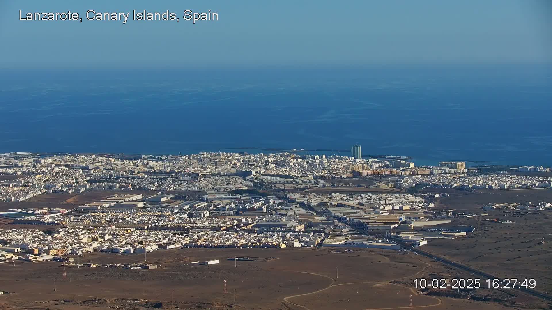 An aerial view shows a coastal city with light-colored buildings stretching to a dark blue ocean under a clear sky.