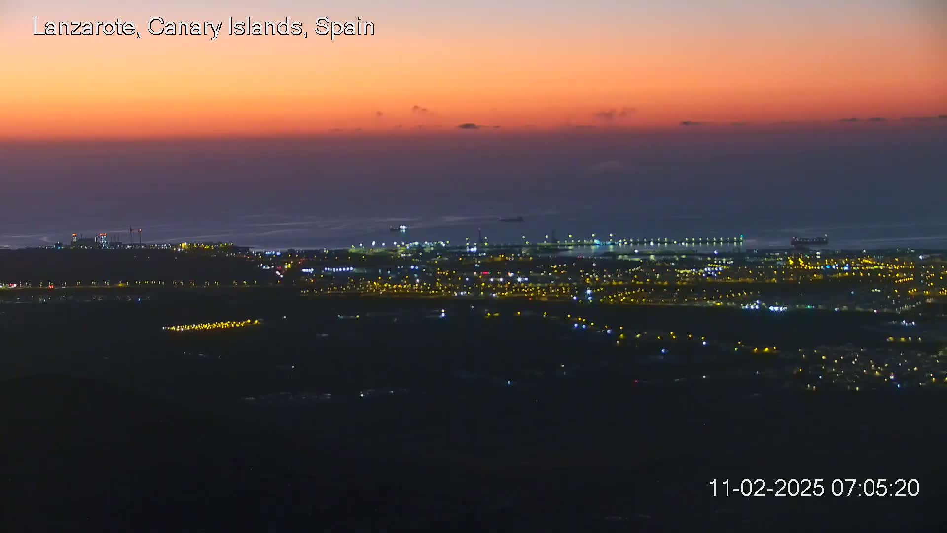 A coastal city at night is illuminated under an orange-pink sunset sky.