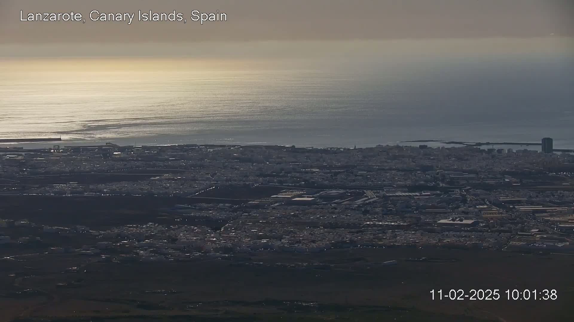 A sprawling coastal city with a dark, mostly overcast sky extends to a calm, reflective ocean under hazy sunlight.