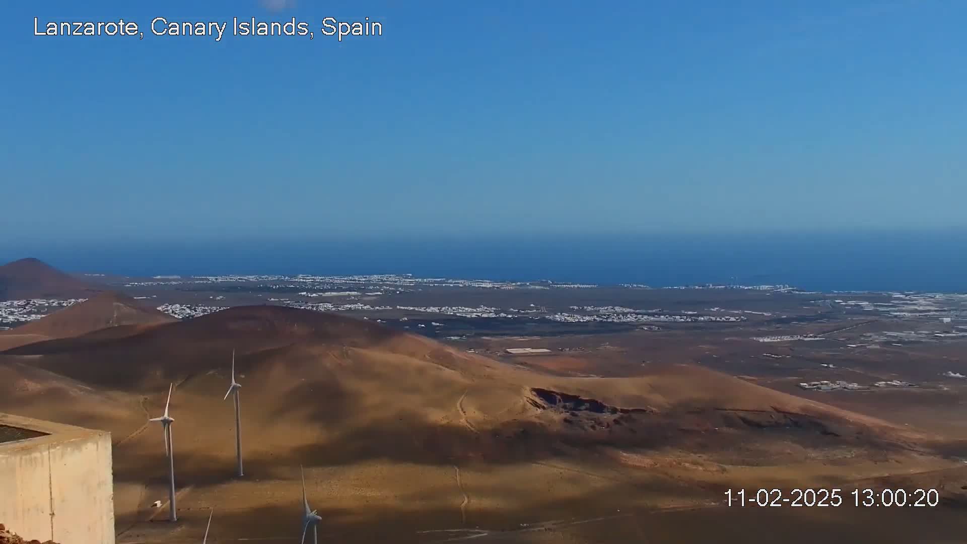 A panoramic view shows a coastal landscape with several wind turbines, low-lying hills, and a distant ocean under a clear blue sky.
