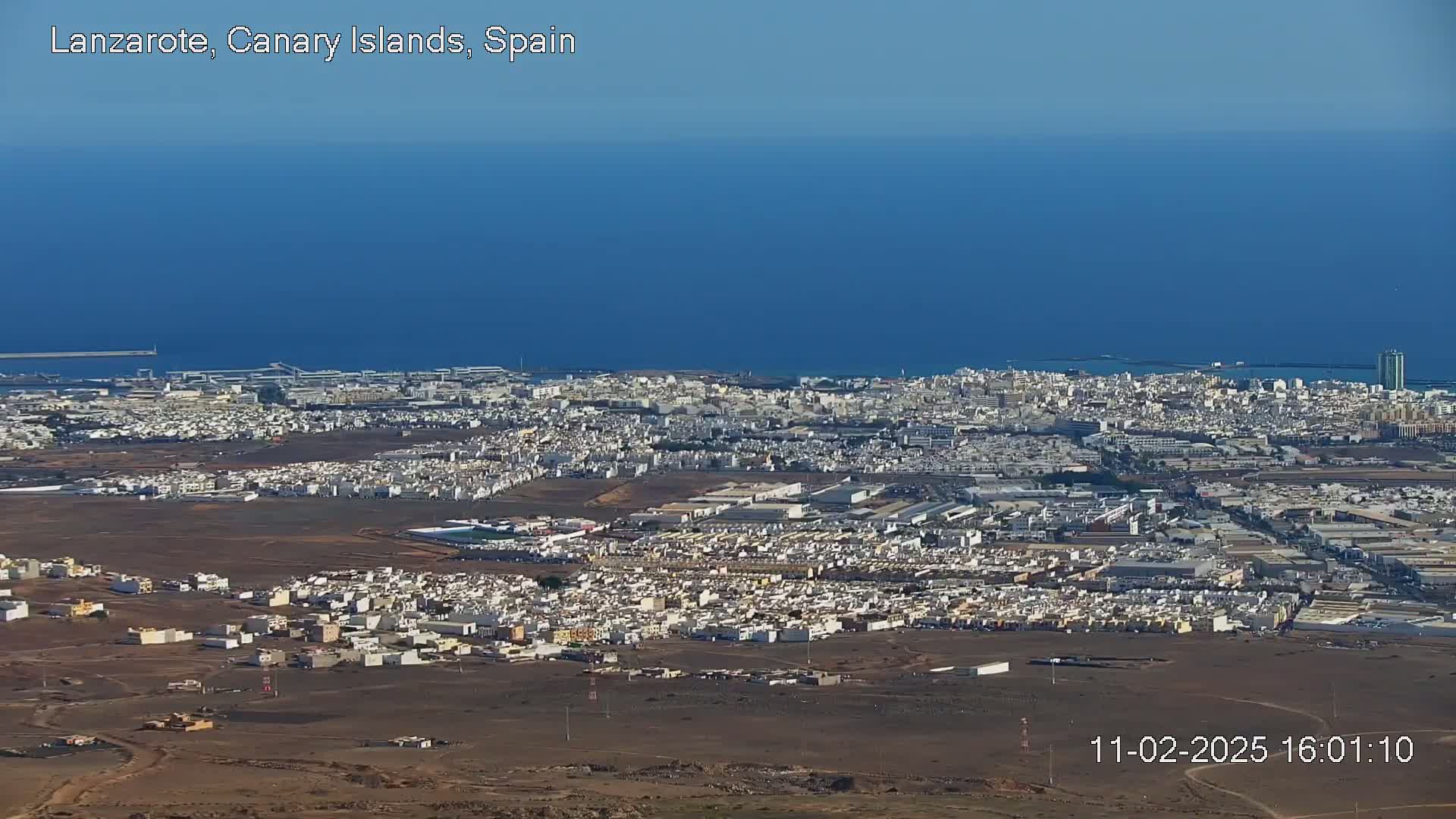 An aerial view shows a sprawling coastal city under a clear blue sky, with a calm, deep blue ocean stretching to the horizon.