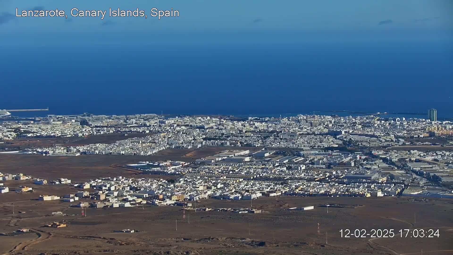 A sprawling coastal city with light-colored buildings extends to a dark blue ocean under a clear sky.