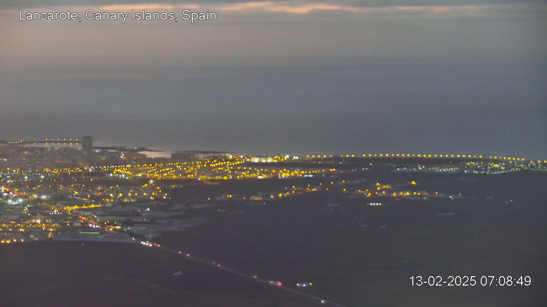 A nighttime, dimly lit coastal city sprawls across a dark landscape under a dusky sky.