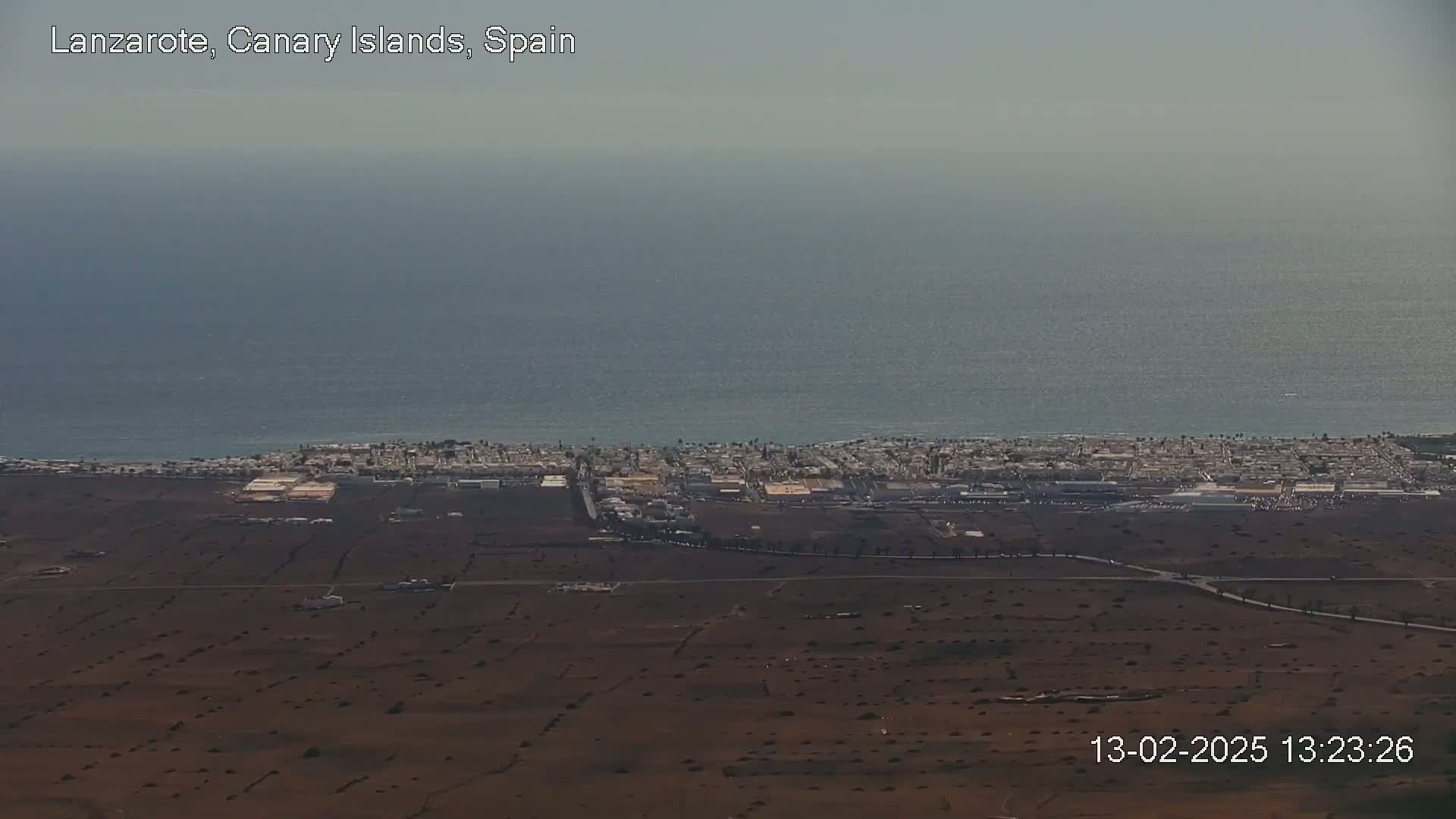 An aerial view shows a coastal town on a flat, dark brown land next to a calm ocean under a hazy sky.