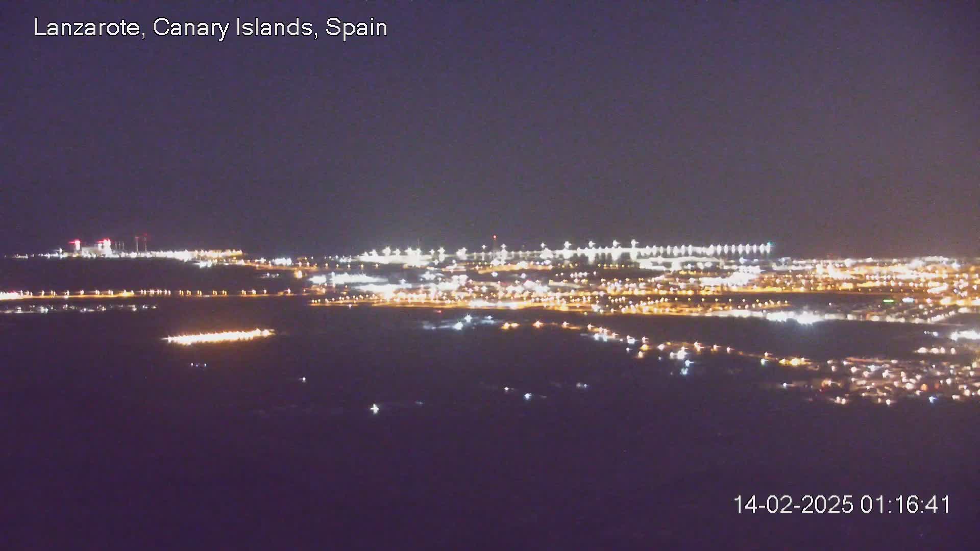 A nighttime, long-range view of a city's illuminated outskirts and airport runway lights stretching across a dark body of water.