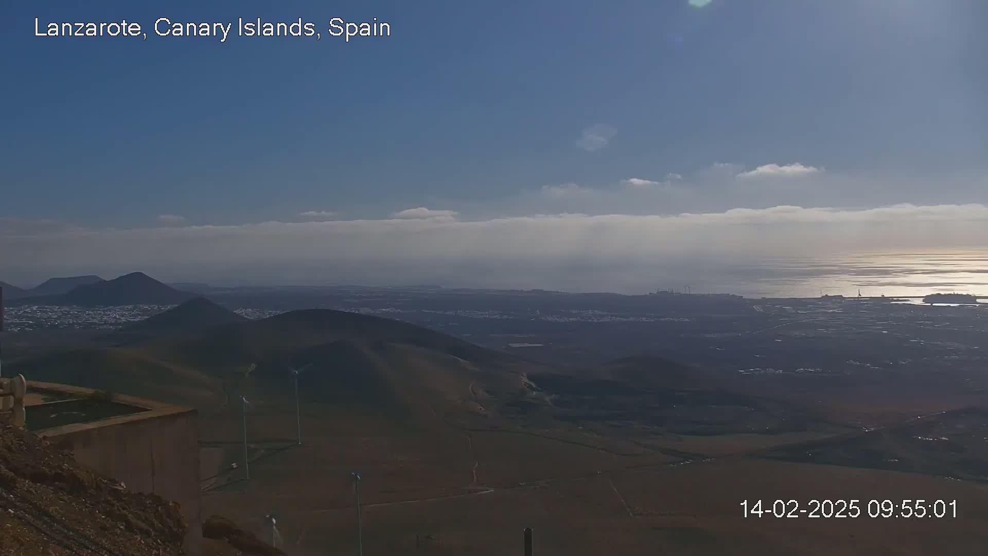 A panoramic view of a volcanic landscape under a mostly clear sky with a few thin clouds shows a distant coastline and ocean under a bright sky.