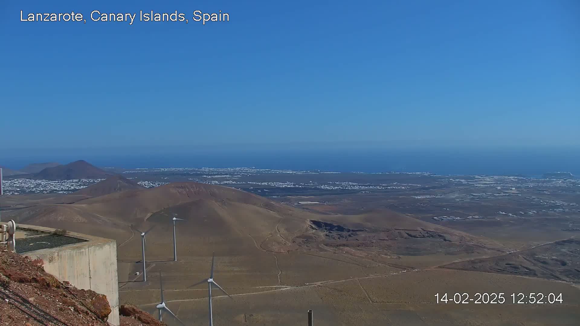 A high-angle view shows a volcanic landscape with wind turbines, a town, and ocean under a clear blue sky.