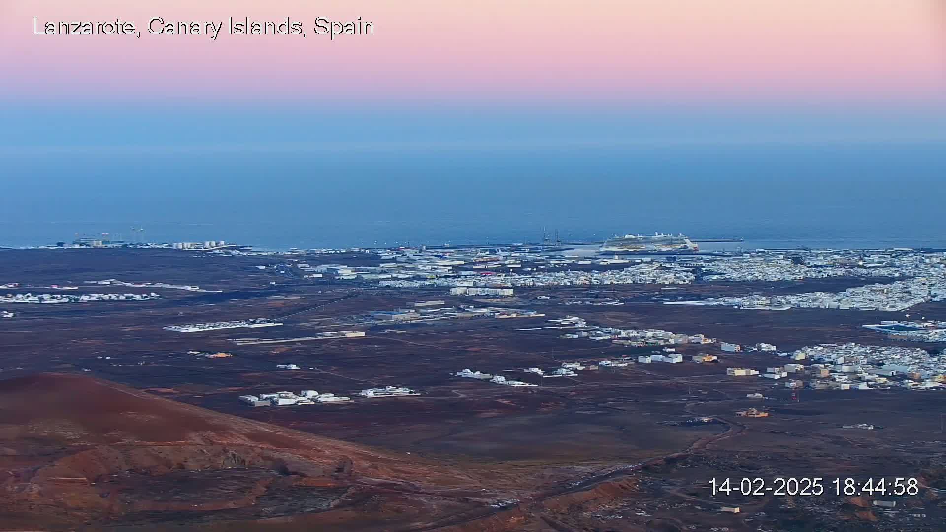An aerial view shows a coastal town with a large cruise ship in the harbor under a soft pink and blue twilight sky.
