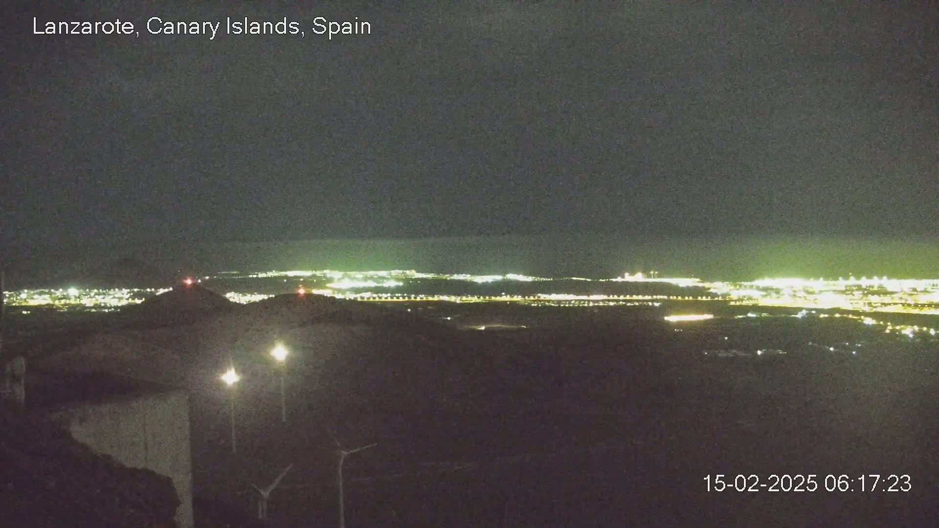 A nighttime, long-range view shows a distant city illuminated against a dark sky, with a closer, dimly lit foreground featuring some structures and what appear to be wind turbines.