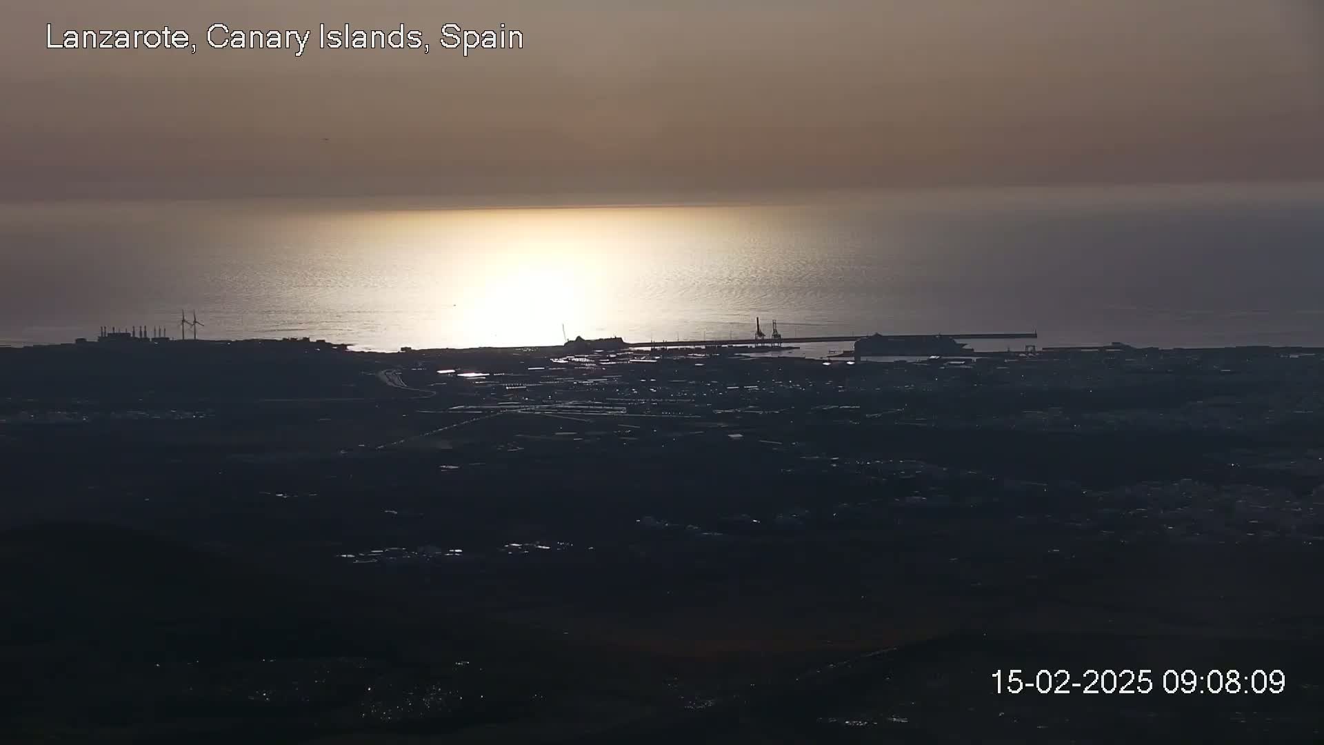 A coastal town at sunset is visible from a high vantage point, showing a port, industrial areas, and a calm sea reflecting the sunlight.