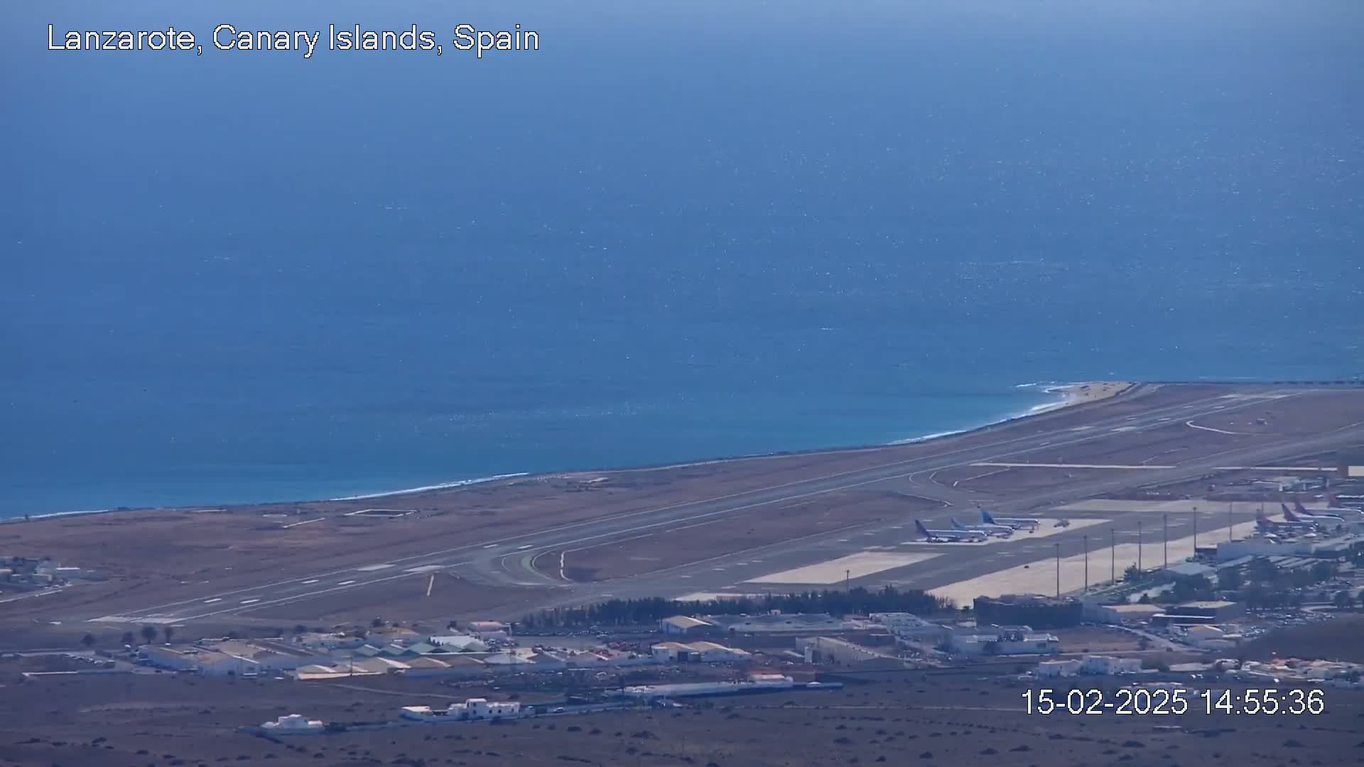 A coastal airport with several airplanes on the tarmac is visible beneath a clear, bright sky, next to a calm ocean.