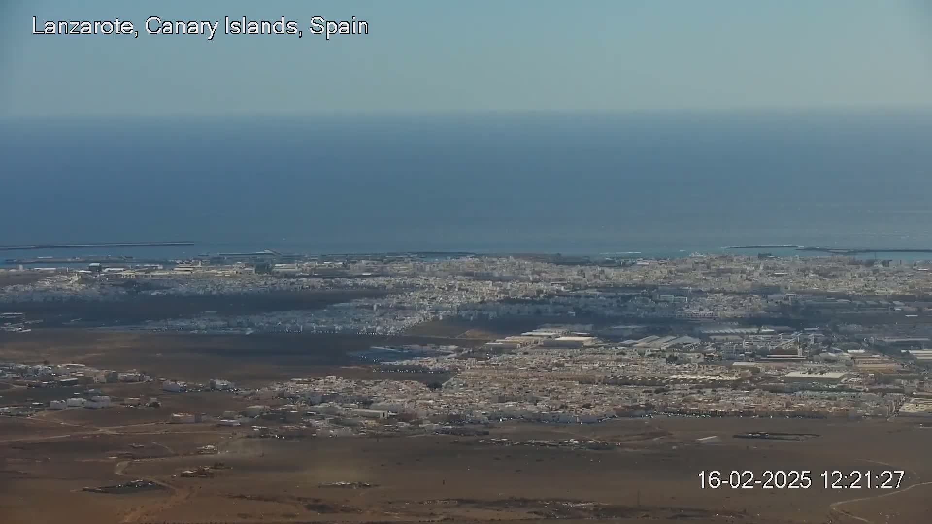 A sprawling coastal city is visible from a high vantage point on a clear, sunny day, with a large body of water extending to the horizon.
