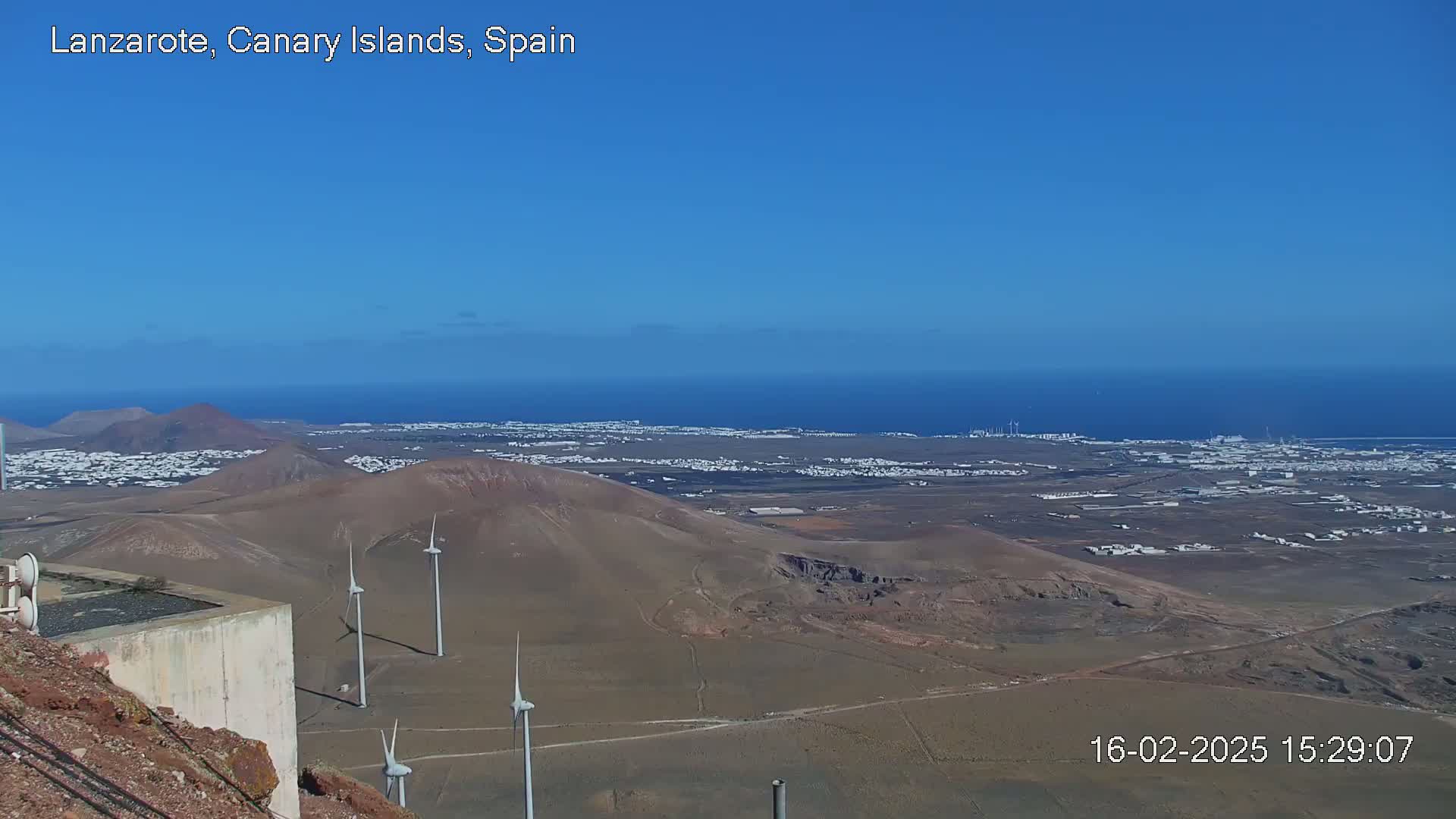 A high-angle view on a clear day shows a volcanic landscape with wind turbines, a town in the distance, and a blue ocean beyond.