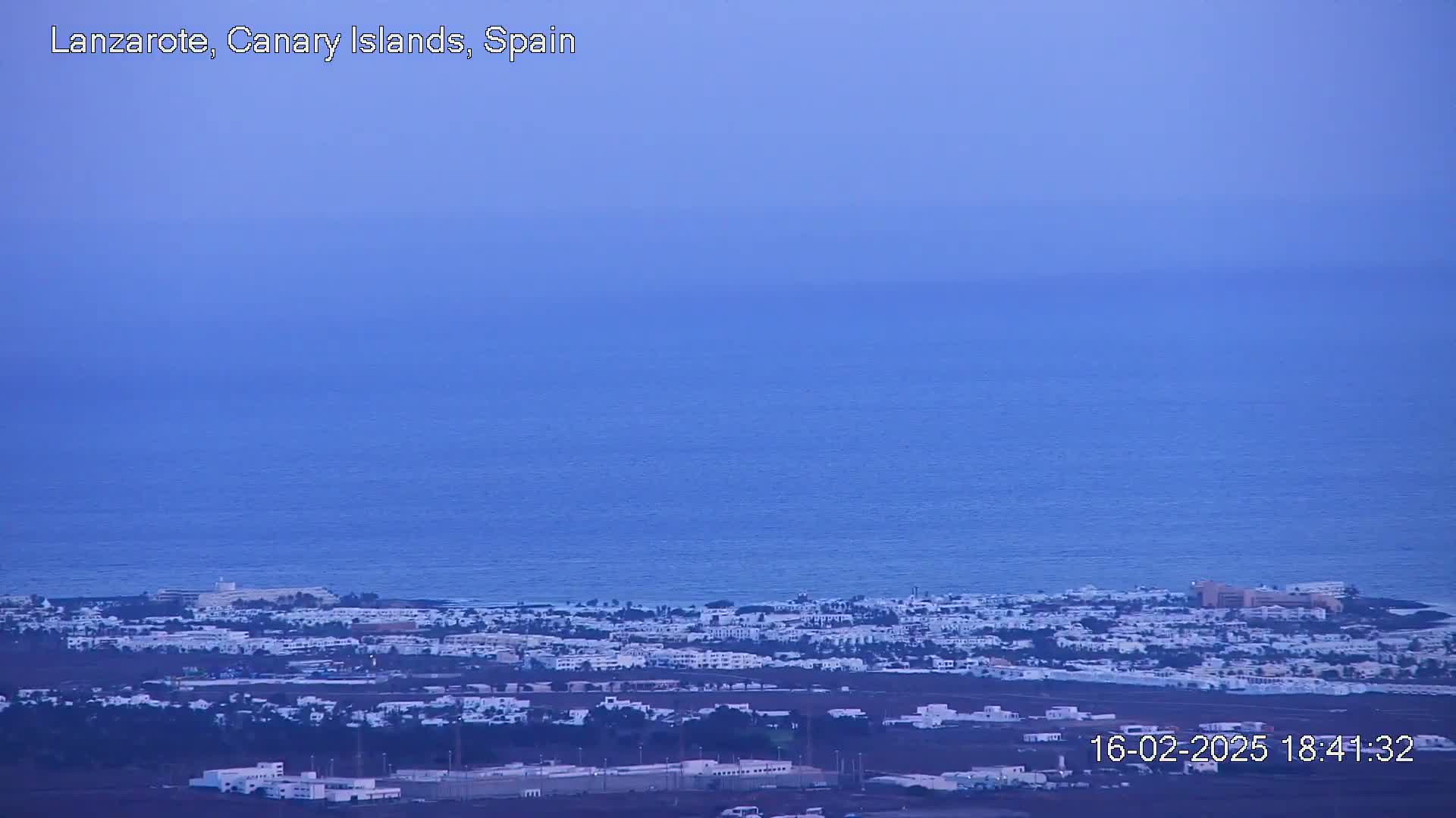 A coastal town with white buildings stretches along a calm, dark blue ocean under a hazy, dusky sky.
