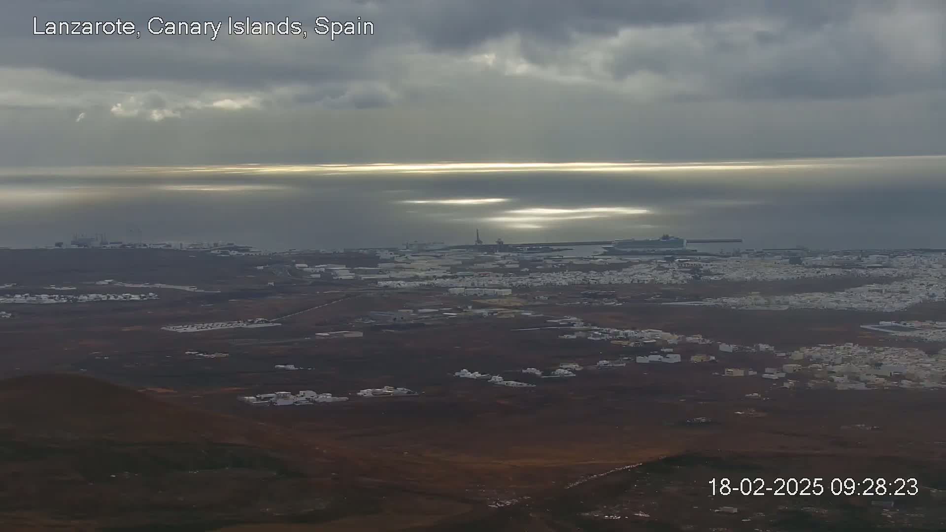 A wide, high-angle view shows a coastal town under a mostly cloudy sky, with the sun reflecting on the water in the distance.