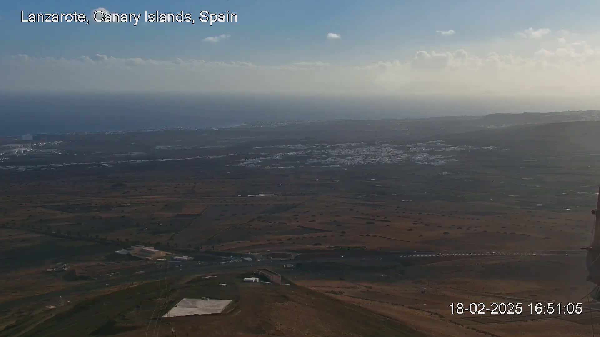 A high-angle, long shot shows a partly cloudy, hazy day overlooking a dry, brown landscape with a town in the distance and ocean beyond.