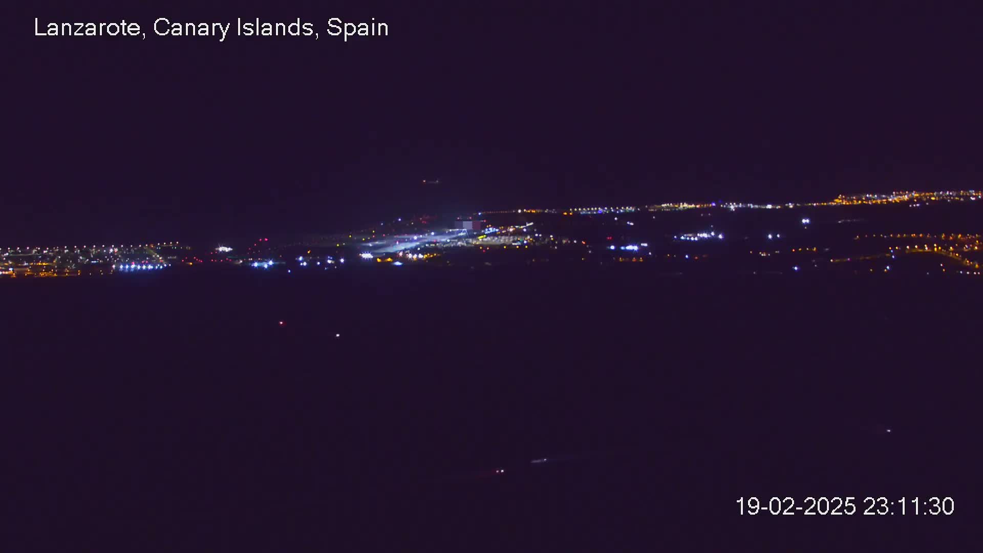 A nighttime long shot of an airport runway and surrounding buildings illuminated by lights under a clear, dark sky.