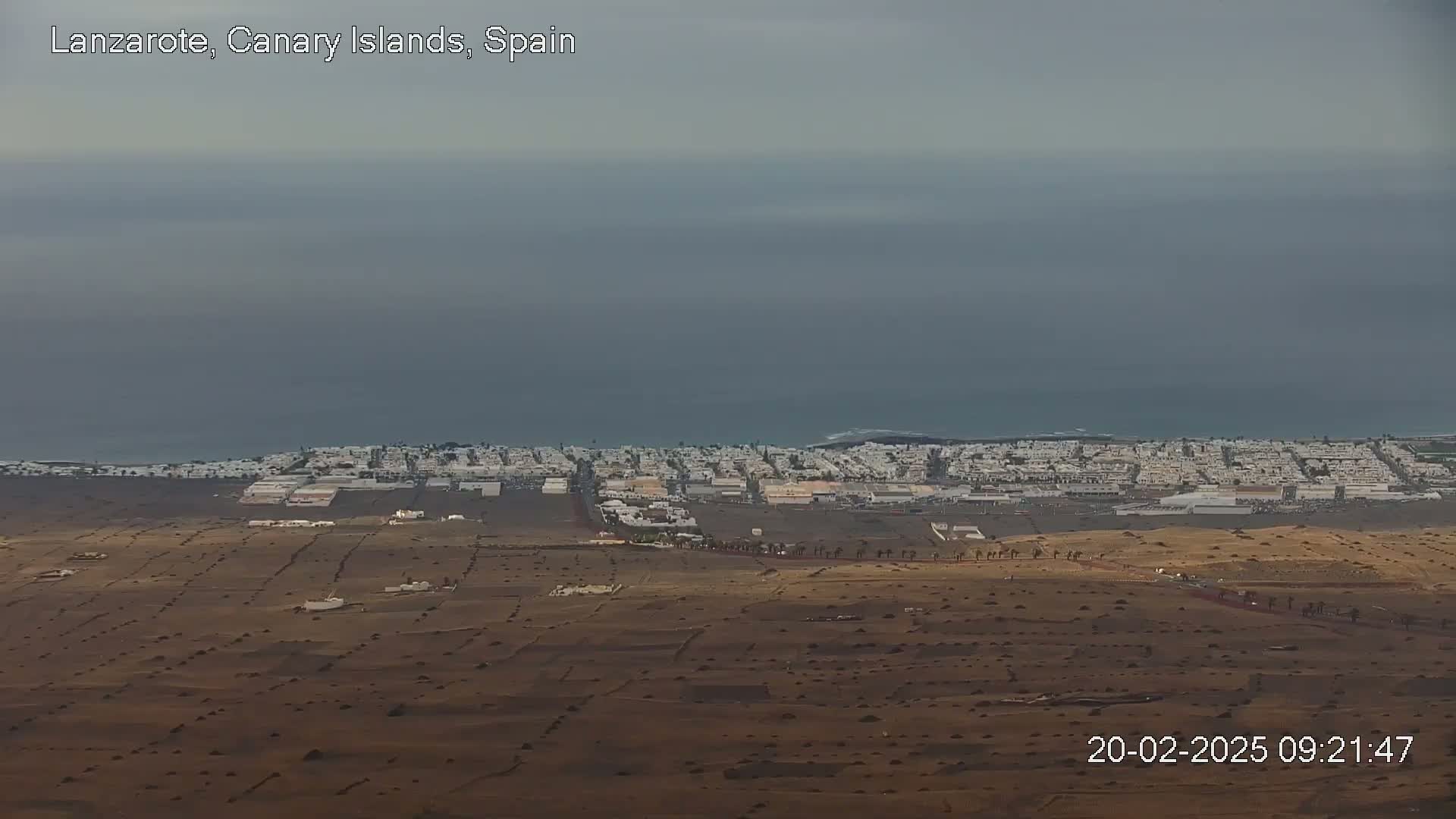 A coastal town with white buildings is visible in the distance under an overcast sky, with dry, brown land stretching out in the foreground.